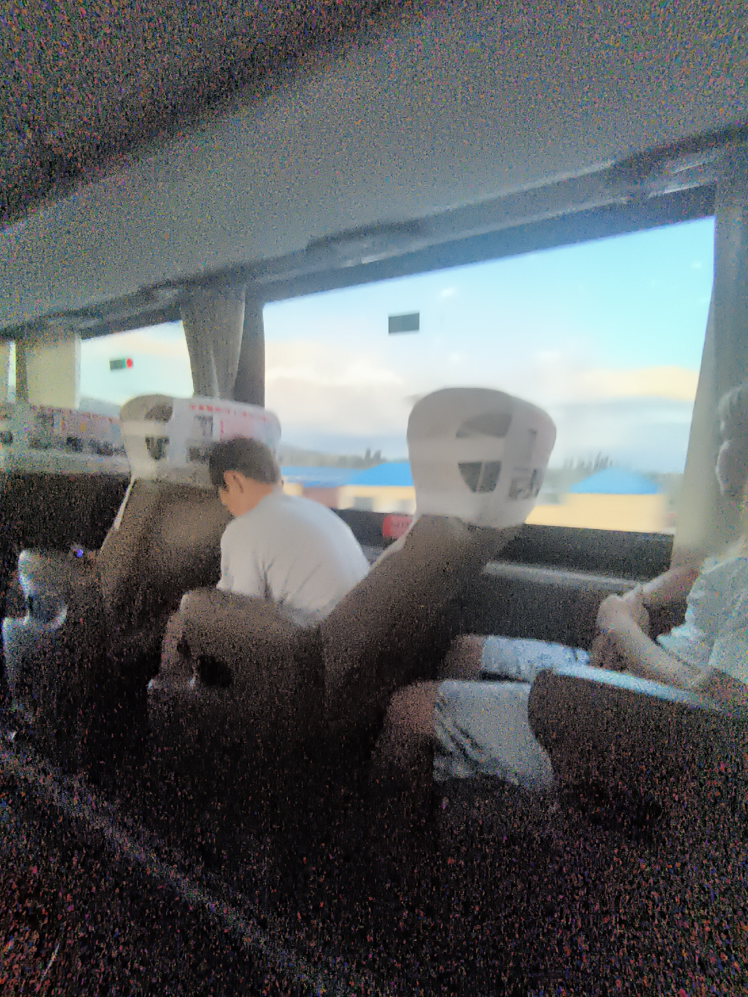 The image, taken from inside a vehicle, shows the interior of a bus or train. A man in a light-colored shirt is seated and facing away from the camera. To the right, another person, only partially visible, is also seated. The background shows a bright sky with clouds, and some blurry distant structures and vegetation, possibly indicating a rural or roadside setting. The image is heavily pixelated, which obscures fine details. The provided location of 果子沟牧场, China, suggests this scene might be in a scenic area known for pastures, though the image itself does not explicitly show any farm animals or open fields. The overall impression is of travel through a landscape.