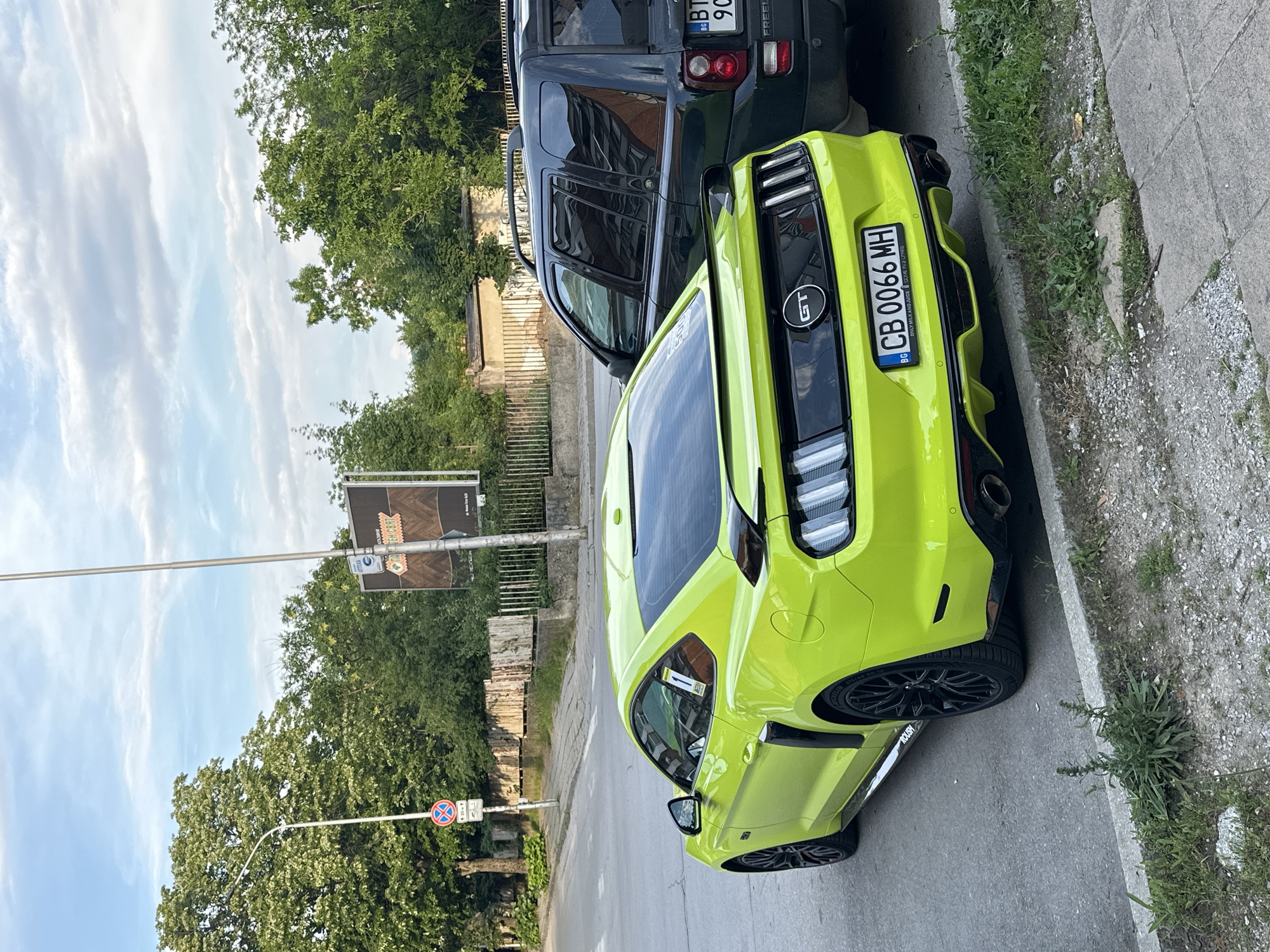 The media file presents an outdoor daytime street scene in Veliko Tarnovo, Bulgaria, dominated by a brightly colored car. The primary subject is a vibrant, almost neon, lime green Ford Mustang GT, seen from a slightly elevated rear perspective, parked on the side of a paved road. Distinctive features of the car include its black wheels, sporty rear diffuser, and a "GT" emblem on the trunk. A sticker on the rear reads "ONLY MILK AND JUICE COME IN 2 LITRES," while its Bulgarian license plate displays "CB 0066 MH." Visible text "ROUSH" on the side suggests it's a performance variant.
Behind the Mustang, a dark-colored SUV is also parked, partially obscured. Its rear shows a Bulgarian license plate beginning with "BG BT 90" and the text "FREEL," likely indicating a Land Rover Freelander model.
The vehicles are parked alongside a concrete sidewalk where weeds and small stones are visible. Lush green trees line the street and form the background, suggesting a verdant area. A metal street pole on the left features a circular 'no stopping' or 'no parking' sign. Another pole further back holds a brown rectangular sign with some visible text, possibly "EH CAT". The sky above is bright with scattered white clouds, indicating clear weather. The overall impression is a typical urban or suburban street environment.