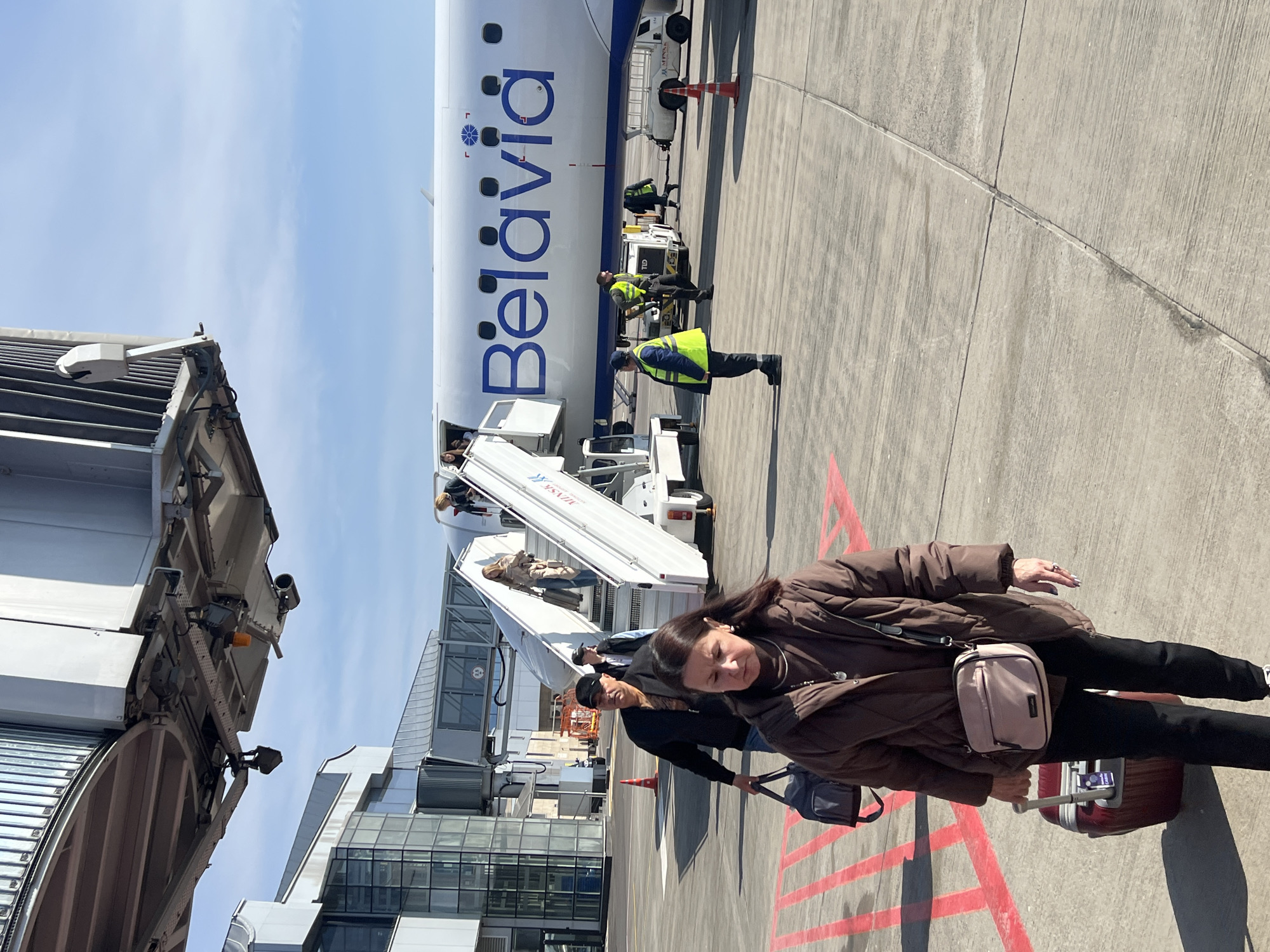 A medium shot taken on a sunny day at the Драчкаўскі сельскі Савет airport in Belarus shows a woman in a brown jacket walking on the tarmac with a red suitcase, in front of a Belavia airplane with workers standing by. The plane is parked near an airport terminal with a jet bridge extended to the aircraft. Passengers are disembarking from the plane using mobile stairs positioned at the front entrance. The tarmac has red directional markings.