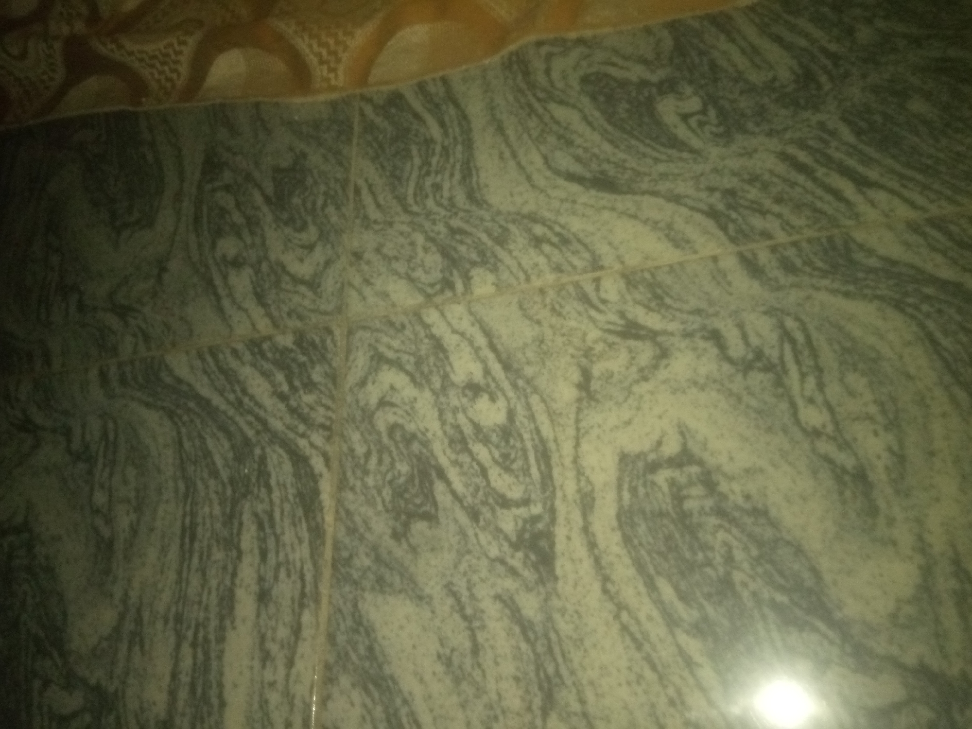 The image shows a close-up, overhead view of marble floor tiles with a swirling, grey and off-white pattern. The tiles are laid in a grid, with visible grout lines. The upper portion of the image shows a glimpse of a patterned rug or carpet. The lighting suggests an indoor setting. There are no people or animals visible. The location is described as Jauro Yinu, Nigeria.