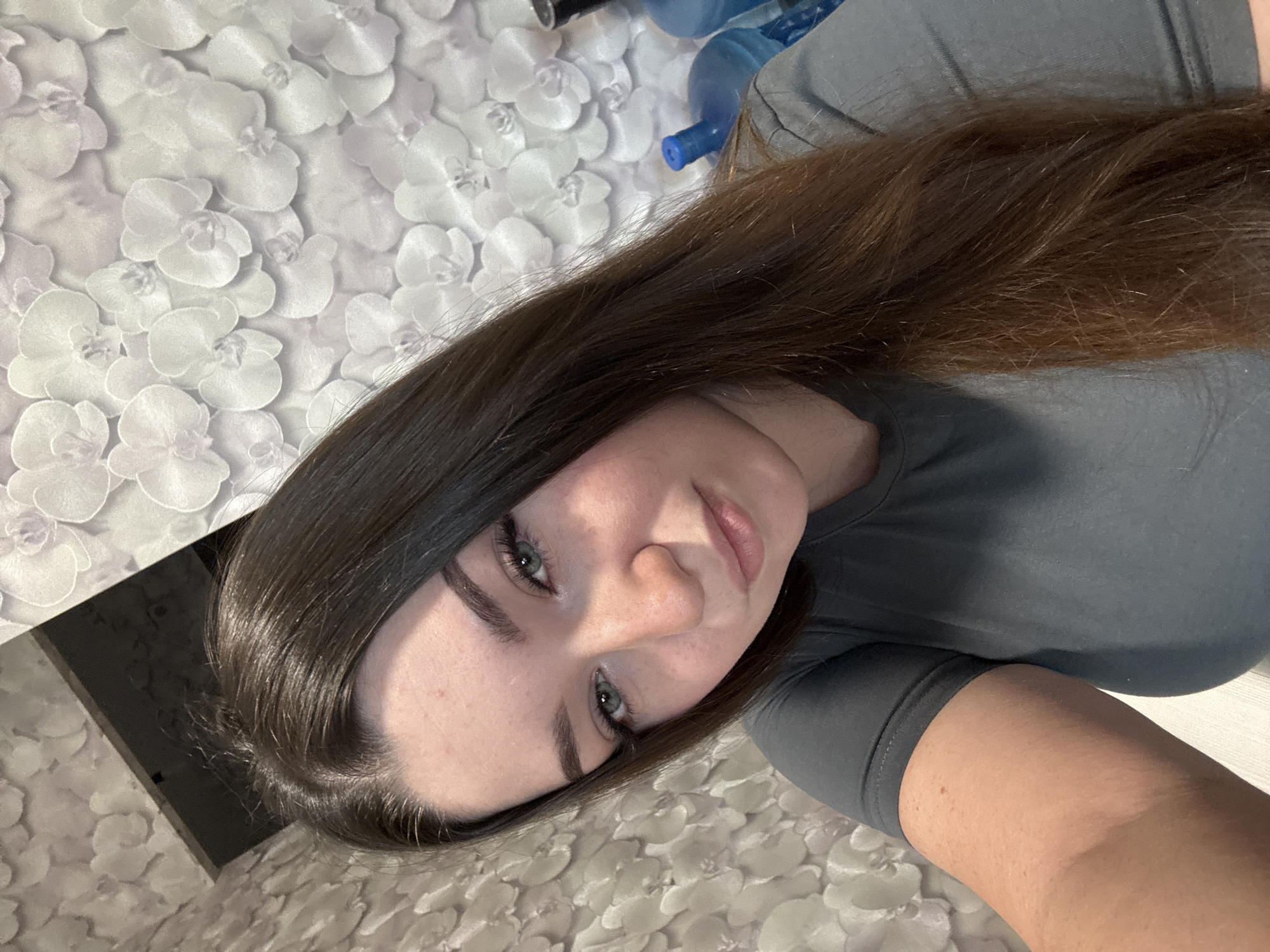 A young woman with long, dark brown hair and green eyes is taking a selfie. She is wearing a grey t-shirt. The background appears to be wallpaper with a floral pattern and a dark rectangular shape in the upper left corner. The scene is indoors, possibly in a room within the city of Zarechnaya (Gamovskoe S/P), Russia. There are large blue water cooler bottles visible behind her to the right.