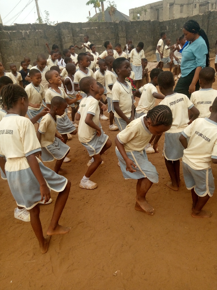 The image depicts an outdoor activity scene in Ukwu-Abwa, Nigeria. Approximately 25-30 young children, primarily wearing cream-colored short-sleeved shirts with "FUTURE LEADER" printed on the back and light blue shorts or skirts with white trim, are actively engaged in movement resembling dancing or exercise. Several children are barefoot, while others wear white sneakers. Their limbs are bent, and expressions suggest exertion or enjoyment. An adult female, wearing a light blue top and dark trousers with braided hair, stands observing the children from the right side of the frame. The activity takes place on reddish-brown compacted earth. In the background, a grey concrete block wall encloses the area, with buildings featuring dark roofs and light walls, along with some sparse vegetation and overhead wires, visible beyond. The sky is overcast.