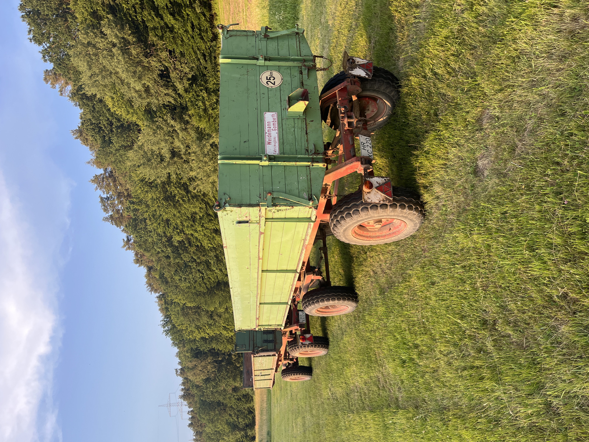 In Wiera, Germany, two green agricultural trailers are parked side-by-side in a grassy field. The trailers are attached to each other and are made of wood with red metal frames and wheels. The larger trailer in the foreground has a "25km" speed limit sticker and a license plate that reads "HR XC 220". The background features a dense forest and a clear blue sky with scattered clouds. The sun is setting, casting a warm glow on the scene. There are no people present in the image.