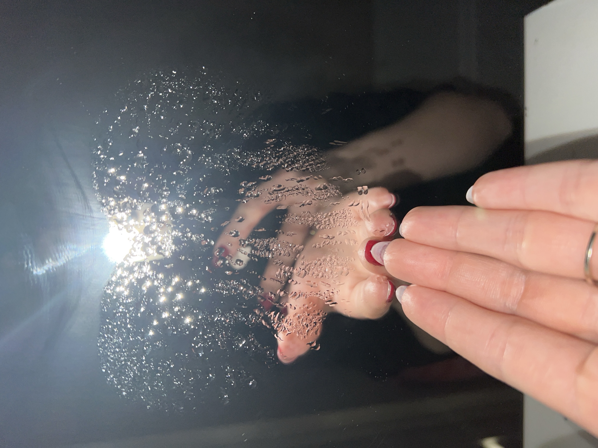A hand with painted fingernails is partially touching a black, reflective surface covered in water droplets. The reflection of the hand is visible in the droplets. A bright light source is also reflected. The location is not specified.