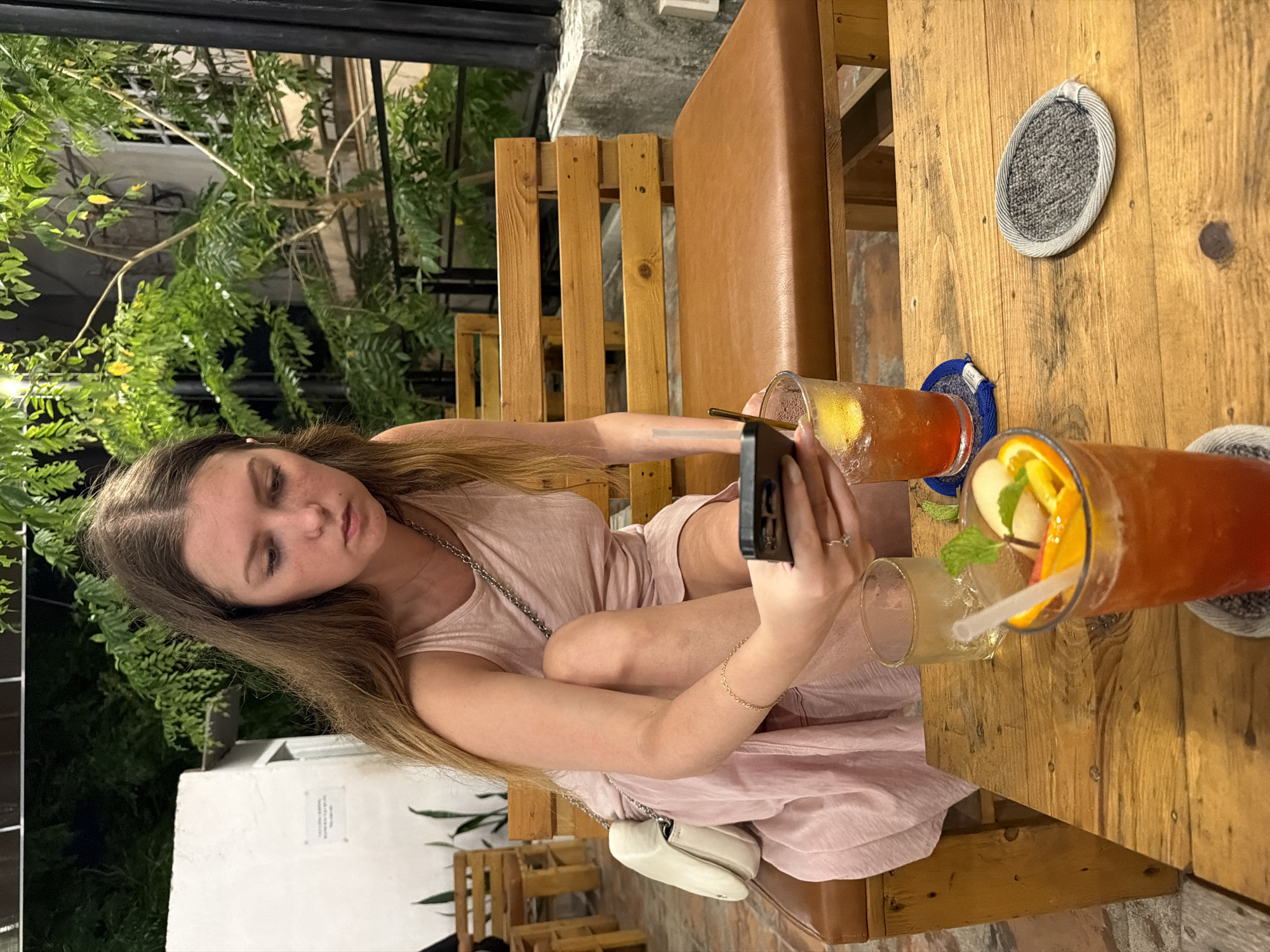 A young Caucasian woman with long, brown hair is seated at a wooden table outdoors, holding a smartphone in her right hand. She is wearing a light pink, sleeveless dress and a white crossbody purse. In front of her are two drinks, one in a tall glass with ice, orange slices, and mint, and another in a shorter glass with ice and a lemon slice. A straw is visible in the tall glass. The table is made of rough-hewn wood. Behind the woman are wooden benches and lush green foliage, suggesting an outdoor seating area, possibly a cafe or restaurant in Bac Nha Trang Ward, Vietnam. The lighting suggests it is evening.