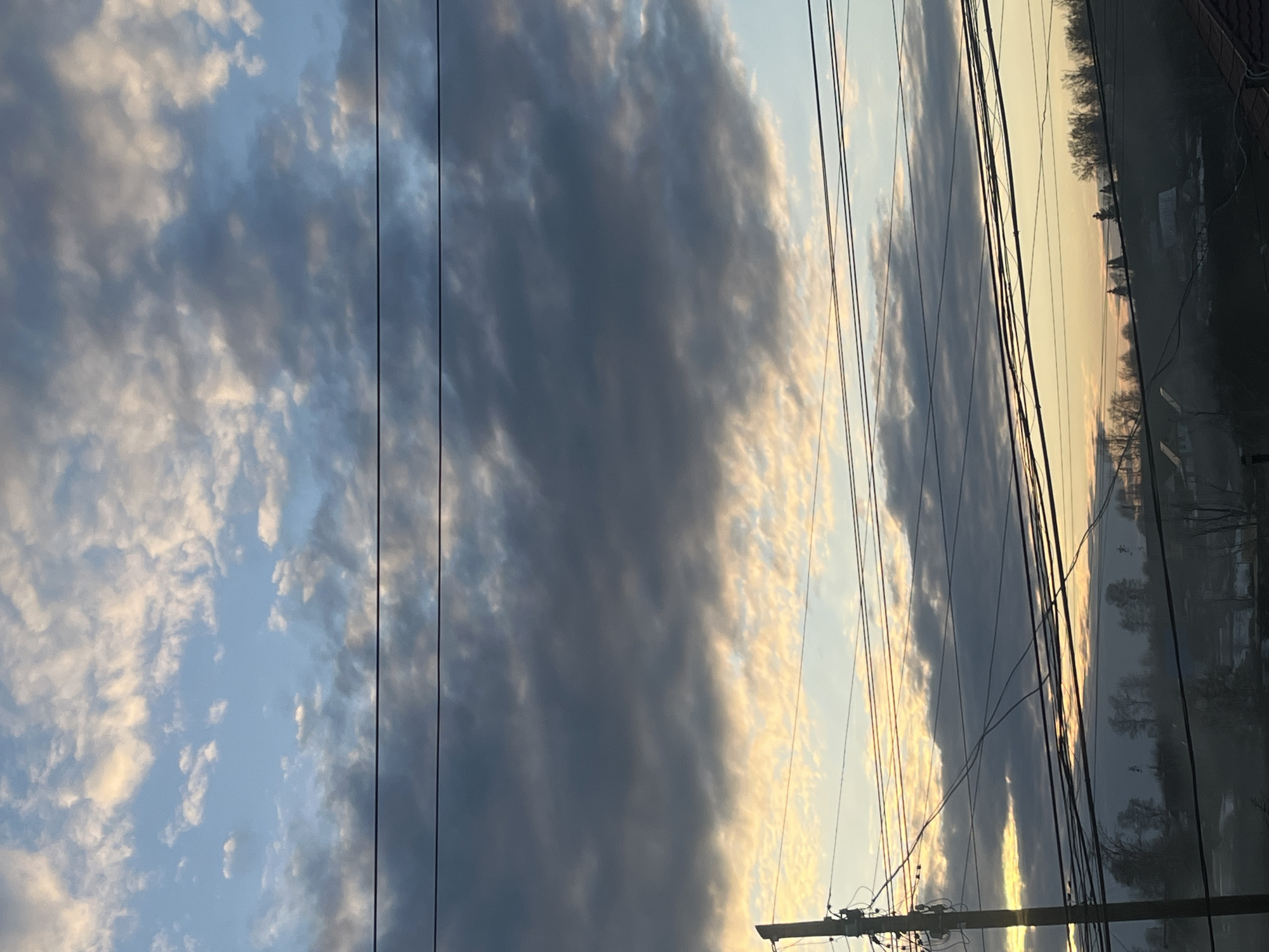 The image captures a landscape at dusk in what appears to be a residential area. High-tension wires crisscross the view of the sky, partially obscuring a bank of dark clouds interspersed with lighter, wispy clouds. The sky has a gradient of light blue to golden hues suggesting a sunset or sunrise. In the background, barely visible through the wires, the outline of trees and buildings hint at the edge of a town, and the location is Фоки, Russia. There are no people immediately visible in the image. The general environment seems calm and still.