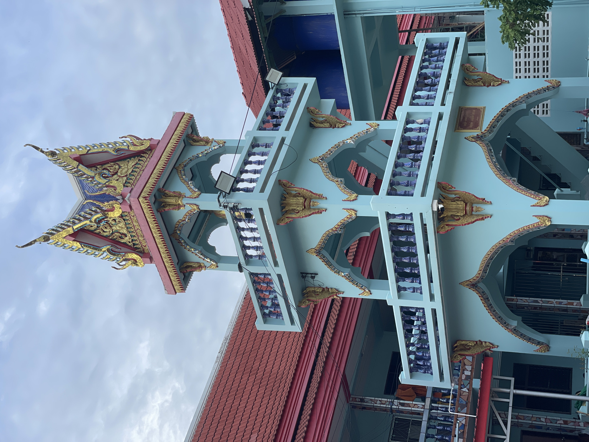 This image depicts a light blue, multi-tiered tower with ornate, golden decorative elements and a colorful, multi-pointed roof, characteristic of Thai temple architecture. The tower is situated in front of a building with a red tiled roof. The sky is overcast, suggesting a cloudy day. The tower features balconies with intricately designed railings and arched openings. The surrounding buildings appear to be part of a larger complex, possibly a temple or a religious institution in Pattaya City, Thailand. No people are visible in the frame. The scene is static, with no discernible actions or interactions. The environment appears to be an outdoor urban or semi-urban setting.