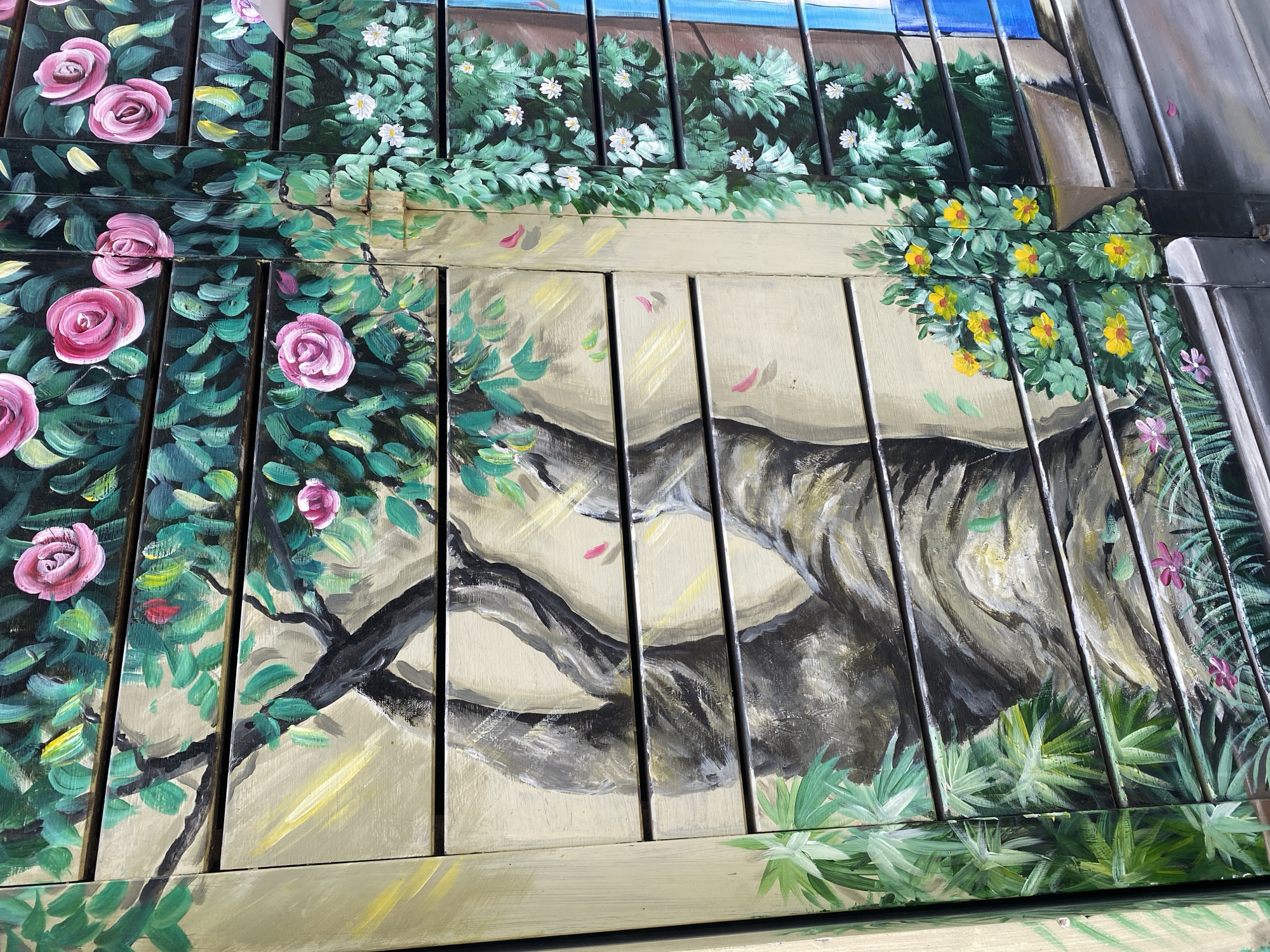 The image depicts a close-up of a mural painted on horizontal slatted panels. The mural features a whimsical garden scene with a large, gnarled tree trunk dominating the left side, rendered in shades of gray and brown with visible brushstrokes. Lush green foliage, painted in various shades of green, surrounds the tree, interspersed with vibrant pink roses and smaller yellow flowers. Some delicate pink petals are scattered across the scene, suggesting a gentle breeze. To the right, a cascade of green vines adorned with tiny white flowers flows downwards, creating a sense of depth. The background is a light, sandy beige, providing a neutral canvas for the artwork. The painting appears to be on an outdoor structure, possibly a shutter or a decorative screen, given the visible slats and the presence of what might be part of a doorway or window frame painted in blue on the far right. The style is illustrative and somewhat impressionistic, with a clear focus on natural elements. This artwork is located in Da Nang, Vietnam, adorning an exterior surface.