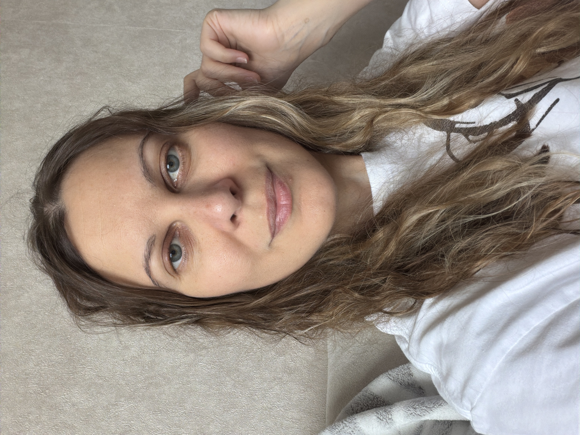 A woman with shoulder-length, wavy blonde and brown hair is lying down, facing the camera. She has fair skin and light blue eyes. She is wearing a white t-shirt, and her hand is raised near her head. The background appears to be a neutral-colored surface, possibly a couch or bed. The location is not discernible as Izhevsk, Russia, or any other city.