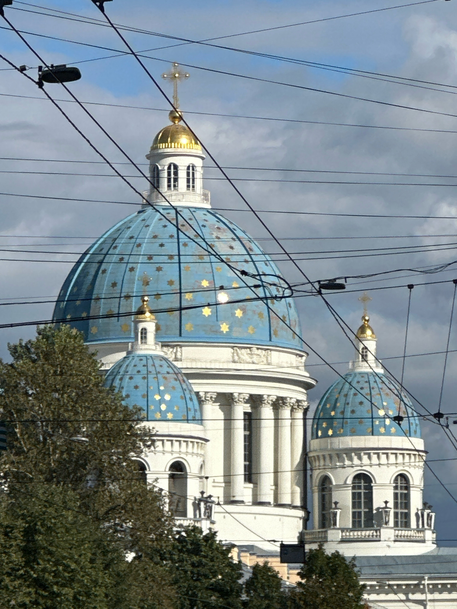 The image shows a close-up view of the blue domes of a Russian Orthodox church, likely the Trinity Cathedral (Troitsky Sobor) in St. Petersburg, Russia, due to its distinctive architecture featuring multiple domes, including a prominent central dome and smaller flanking domes. The domes are adorned with gold stars. The church is partially obscured by a dense network of black electrical wires crisscrossing the foreground, common in urban environments. In the lower left portion of the image, lush green trees are visible. The sky above is a mix of blue and gray clouds. The architecture is grand and ornate, with white columns and arched windows visible on the church's structure. The sunlight illuminates the gold stars on the domes, making them stand out against the blue.
