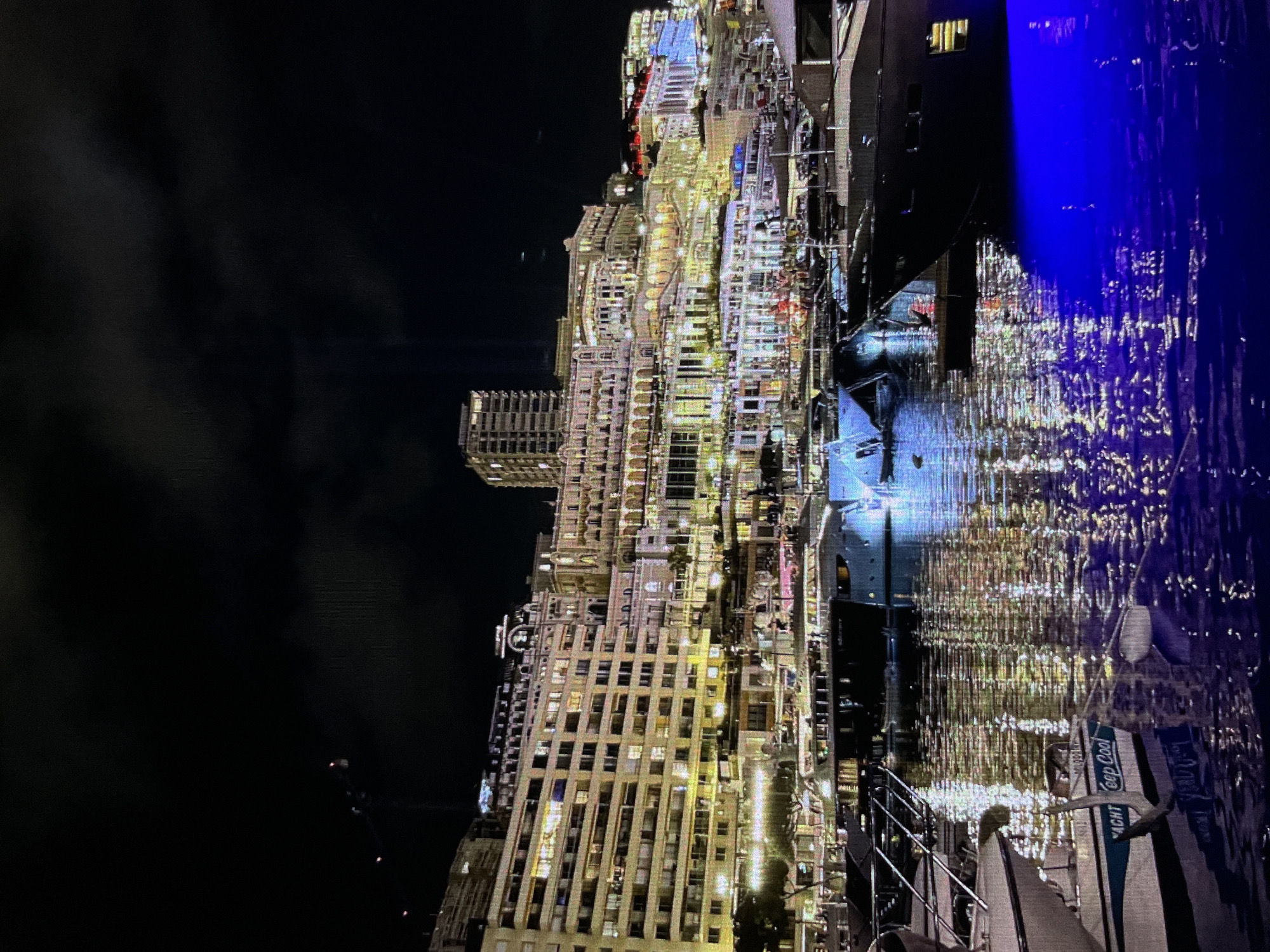 The image captures a nighttime view of a harbor in what appears to be Monaco. A large, illuminated cityscape with numerous buildings and hotels rises behind the harbor. The buildings are a mix of architectural styles, with many featuring extensive lighting, creating a warm glow that reflects on the water. Several yachts of varying sizes are docked in the foreground, their shapes and lights visible against the dark water. The water itself is illuminated by the city lights and also by a prominent blue light source, possibly from one of the boats or underwater lighting, creating a dramatic effect on the surface. In the upper left portion of the image, a construction crane is visible against the dark night sky, suggesting ongoing development in the area. The overall scene conveys a sense of luxury and activity at night in a vibrant coastal city.