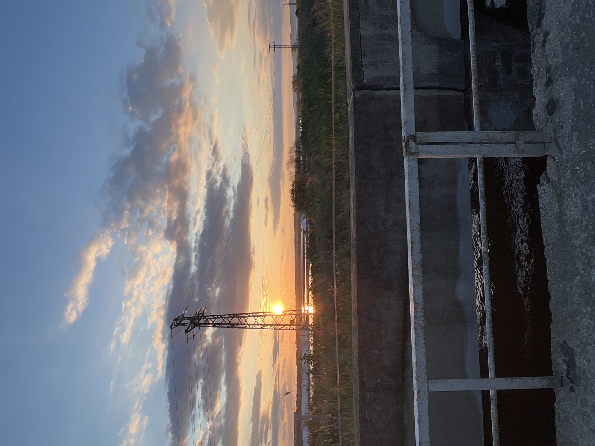 The image shows a landscape taken from what appears to be a bridge or elevated structure, potentially in Burgas, Bulgaria. The foreground includes a concrete structure with metal railings and openings. Through the railings, there is a view of a sunset or sunrise. A transmission tower stands in the middle ground, silhouetted against the light. The sky is filled with clouds tinged with orange and yellow hues from the setting sun. Beyond the tower, there is a stretch of land with vegetation.