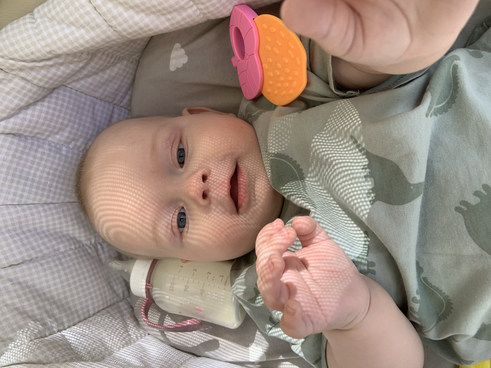 This is a close-up, high-angle shot of a baby lying in a crib. The baby has fair skin, blue eyes, and blonde hair. Sunlight casts a distinct grid pattern across the baby's face. In the foreground, the baby's right hand is visible, with its fingers slightly curled. To the left of the baby is a baby bottle filled with a pale liquid, with measurement markings in milliliters and ounces visible. The bottle has a pink strap attached. Above the baby's head, a colorful teether toy with orange and pink sections is visible.
The baby is wearing a pale green onesie with a repeating pattern of gray dinosaur-like creatures. The crib appears to be lined with beige and white checkered fabric, and a gray blanket with a white cloud imprint is visible in the background.
The scene suggests a moment of rest or play for the baby. The sunlight and the baby's relaxed pose indicate it's likely daytime. The presence of the baby bottle and teether points to the baby's age and needs. The overall mood is peaceful and intimate.
Visible text on the bottle reads "MADE IN EU - BPA Free". The location context is Kyiv, Ukraine.
