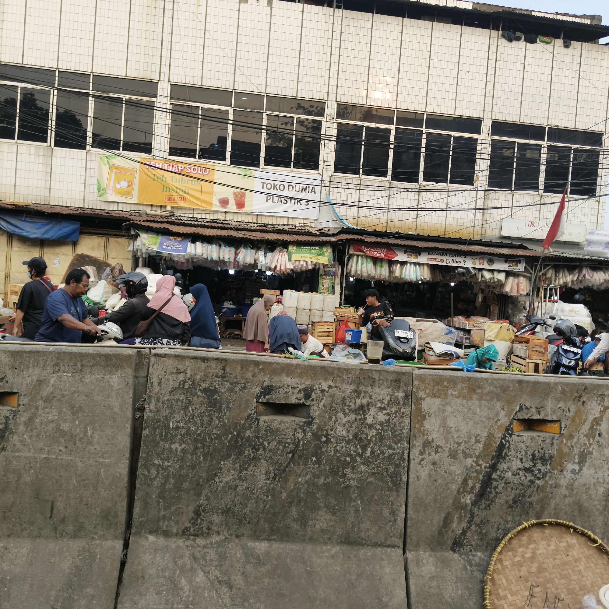 The image depicts a street scene in South Jakarta, Indonesia, possibly near a market or commercial area. In the foreground, a series of weathered concrete barriers form a divider. Behind the barriers, a row of shops or stalls is visible under a corrugated metal roof. The stalls display a variety of goods, including plastic containers, packaged food items, and what appear to be textiles or bags. Several people are present, some interacting with each other and others tending to their merchandise.
In the middle ground, a group of individuals is gathered, some sitting on motorcycles. One man in a blue shirt is holding onto the handlebars of a scooter, looking forward. Another man wearing a black shirt and a blue mask is standing behind him. A person wearing a black helmet and a dark jacket is seated on a motorcycle, facing away from the camera. A woman in a pink hijab is seated behind the motorcyclist, and another woman in a blue hijab is seated to her right.
In the background, a multi-story building with white tiled facade and rows of dark-tinted windows is visible. Banners are displayed on the building's lower level, one of which is yellow and features text in Indonesian, possibly advertising "Teh Tiap Solo" and "Toko Dunia Plastik". Another banner, white with red lettering, reads "Aladdin Frozen". A red and white Indonesian flag is flying from a pole near the top right of the building. The overall environment appears to be a bustling commercial area with elements of both permanent structures and temporary market stalls. The lighting suggests it could be daytime, possibly late afternoon, given the shadows.