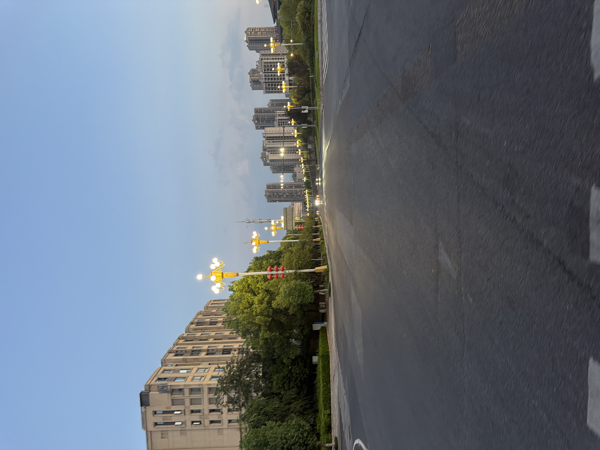 This is a wide-angle, eye-level shot of a street in Jiangjin, China, taken at dusk. The sky is a clear, pale blue, transitioning to a lighter hue towards the horizon, with some faint clouds visible. The street is paved with asphalt and appears wet, reflecting the glow of streetlights. Pedestrian crosswalk markings are visible on the road.
On the left side of the street, a multi-story residential building with a beige facade and numerous windows lines the sidewalk. Lush green trees and manicured hedges are situated between the building and the road. Several ornate, yellow streetlights with multiple bulbs illuminate the scene, some also featuring red lantern decorations.
To the right, the street stretches into the distance, lined with more of the same yellow streetlights. Further down, a row of modern, high-rise apartment buildings dominates the skyline. The lights from these buildings are visible, suggesting an urban environment. A few vehicles with their headlights on can be seen in the distance, indicating light traffic. The overall impression is that of a quiet, early evening in a developed urban area.