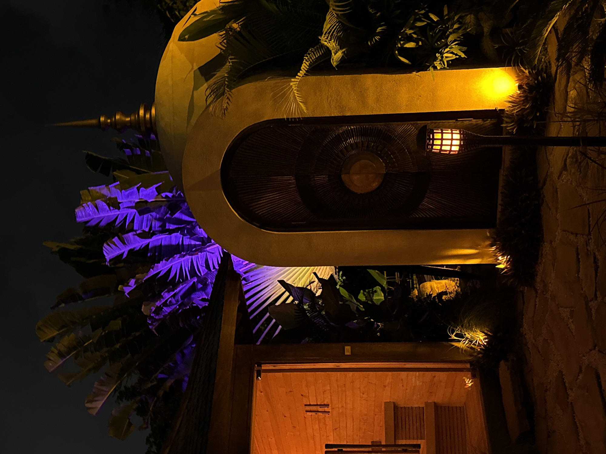 At night, an architectural feature is illuminated with yellow light in Mengwi, Indonesia. The structure is situated amongst dense vegetation. A rounded dome with a spire is decorated with purple lighting. Under the dome, there is a covered area with a ceiling fan, lit by a lantern-style light. Below, a partially visible wooden structure suggests a room or entry. Stone texture is seen on a wall to the right.