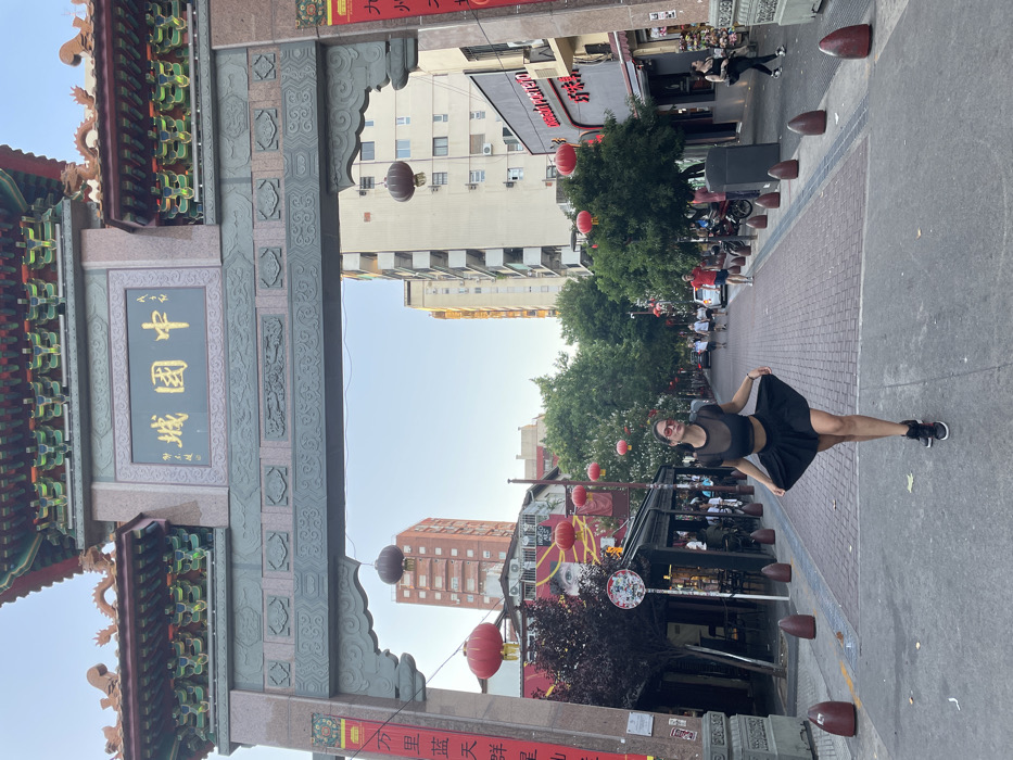 A young woman in a black outfit and sunglasses stands on a brick street under a Chinese archway entrance. The architecture includes ornate carvings and Chinese characters. Buildings line the street, adorned with red lanterns and trees. People are seen further down the street, suggesting an active urban area. The sky is clear and bright. The scene evokes a sense of cultural immersion within a Chinatown setting.
