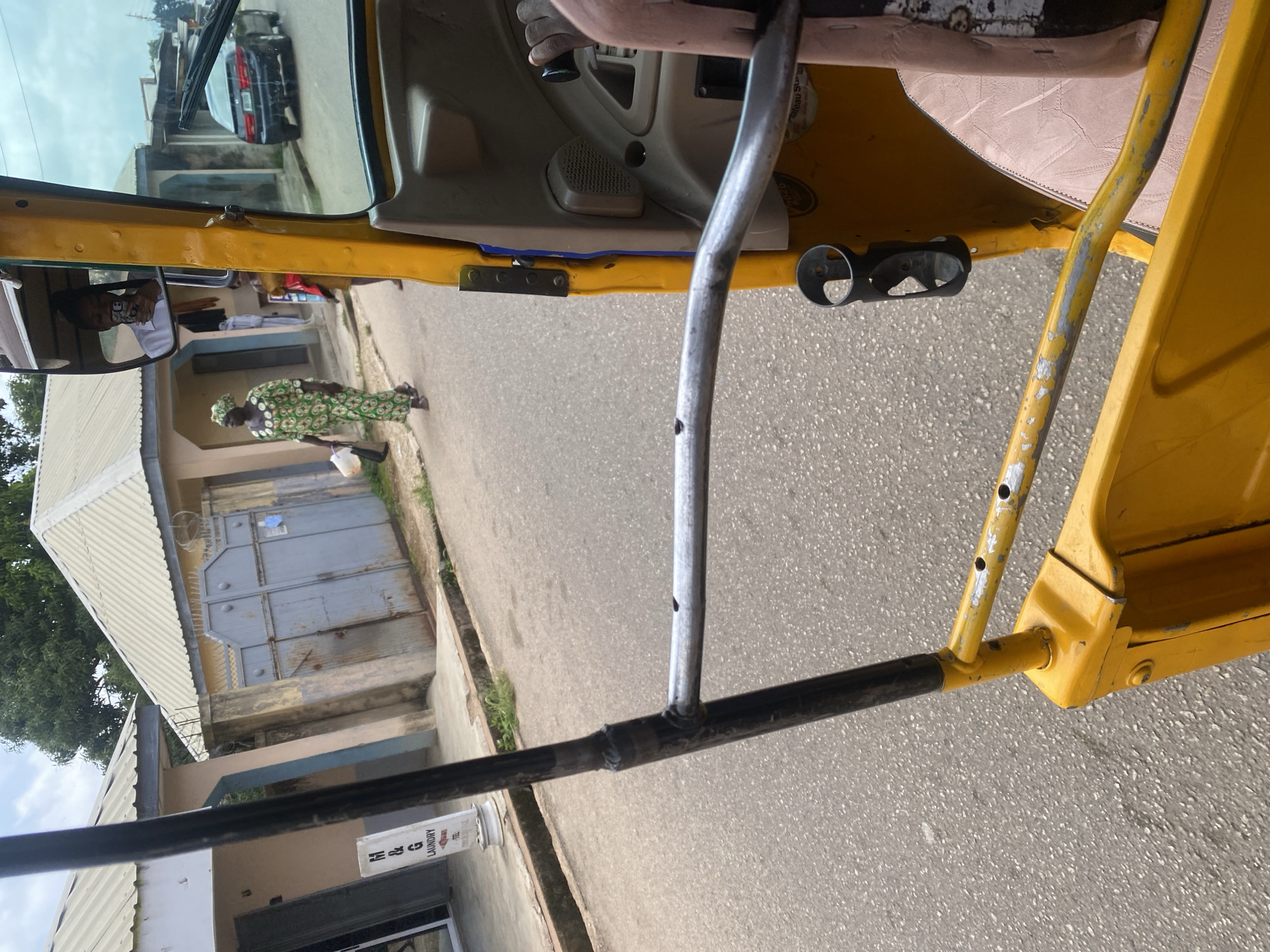 An outdoor, eye-level, medium shot captures a scene from inside a yellow auto-rickshaw, looking forward. The auto-rickshaw has a weathered exterior, with visible metal structures and some paint chips. In the foreground, a black metal bar extends horizontally across the frame. A dark holder for a cup or bottle is attached to the yellow frame. To the right, part of a light pink seat is visible.
Beyond the auto-rickshaw, a street runs through a residential area in Jos, Nigeria. Buildings with corrugated metal roofs line the street. One building on the left has a sign that reads "M & G LAUNDRY." A large grey metal gate is visible on the right side of the street.
A woman in a green and white patterned traditional dress walks away from the camera on the sidewalk. Her head is covered by a matching headscarf. She carries a white bucket and a dark bag. Behind her, another person in a red top is partially visible. In the background, a dark-colored car is parked on the right side of the street. The sky is overcast, suggesting a cloudy day. A reflection in the auto-rickshaw's side mirror shows a person taking a selfie.