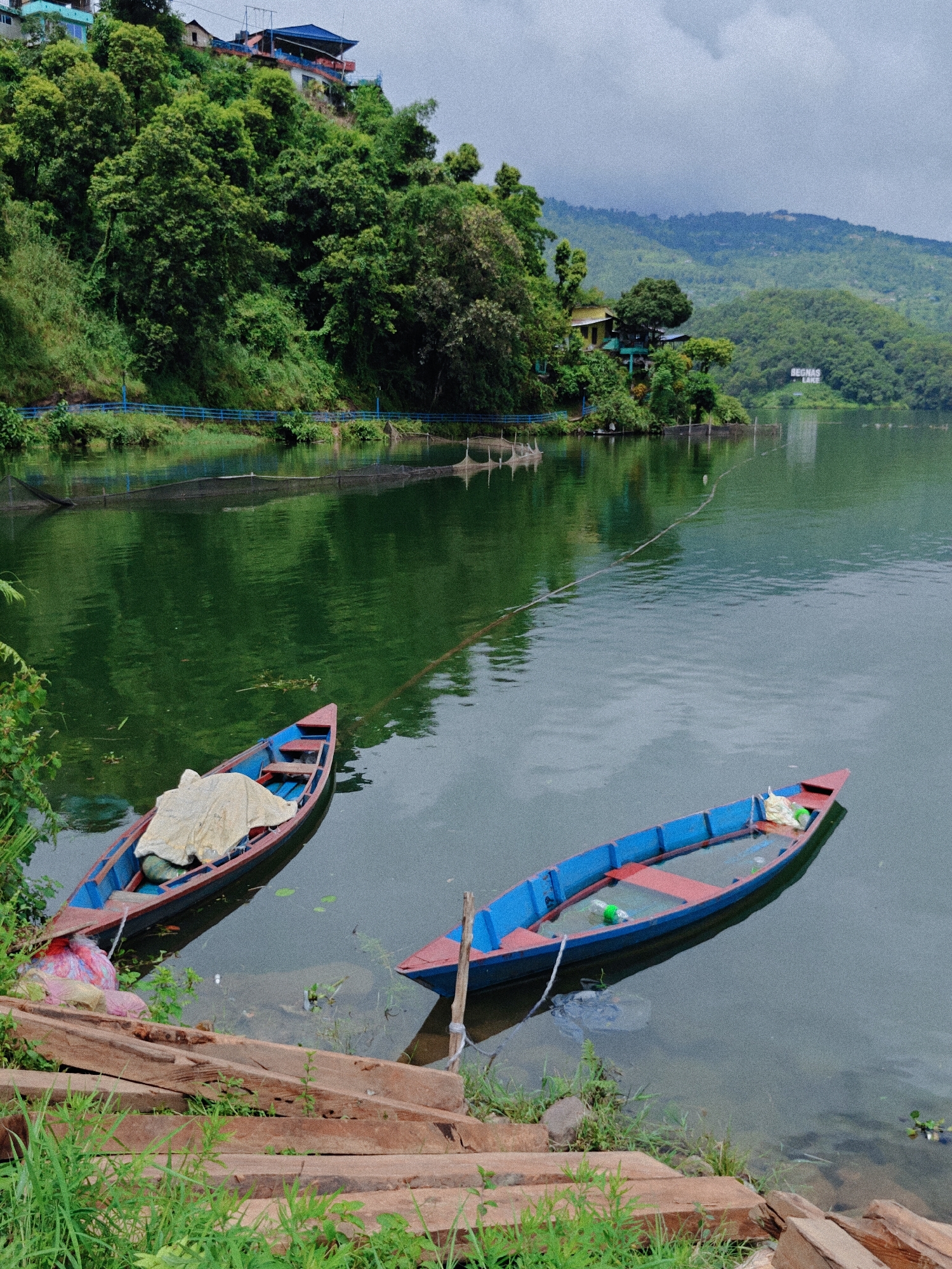 This is a high-angle, medium shot of Begnas Lake in Pokhara, Nepal. Two traditional wooden boats are in the foreground, moored near the shore. The boat on the left is mostly covered with a tan tarp, while the boat on the right is partially submerged and contains some plastic bottles and other debris. The lake's water is a deep green, reflecting the lush, tree-covered hills that rise steeply behind it. In the distance, houses and structures are visible nestled amongst the trees on the hillside, and a sign reading "BEGNAS LAKE" can be seen further down the shore. The sky is overcast with a light haze. The shore in the foreground is lined with wooden planks and tall grass. There are no people visible in the image.