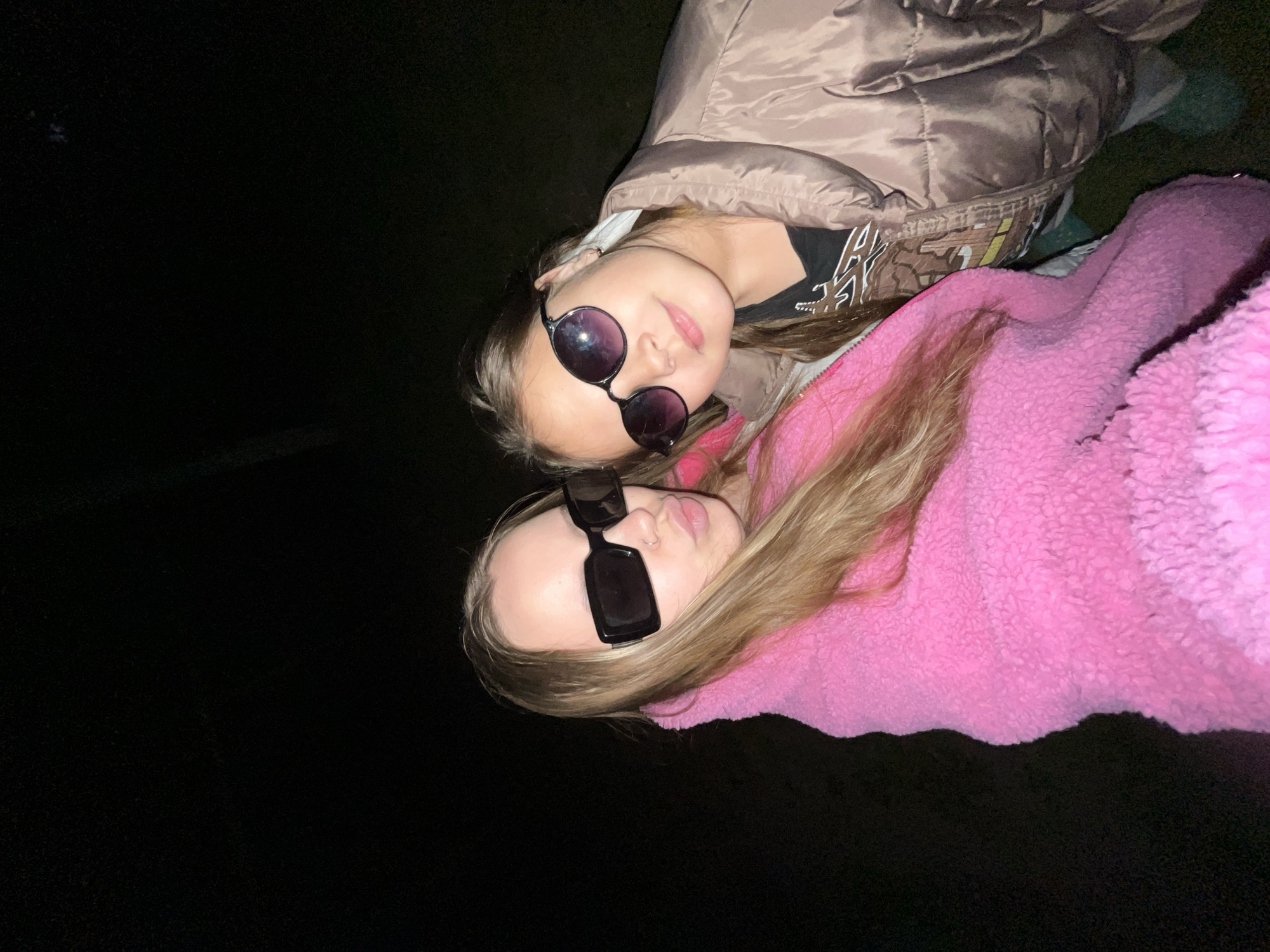 Two young girls are lying down close together in a dark environment. The girl in the foreground wears a pink, textured jacket and black, rectangular sunglasses. Her blonde hair is partially visible. The girl behind her wears a light brown, quilted jacket, a black t-shirt with white letters, and round, dark-lensed sunglasses. The background is predominantly black, suggesting a night setting. The location in the city of Novopokrovskaya, Russia, is not determinable from the visual information provided.