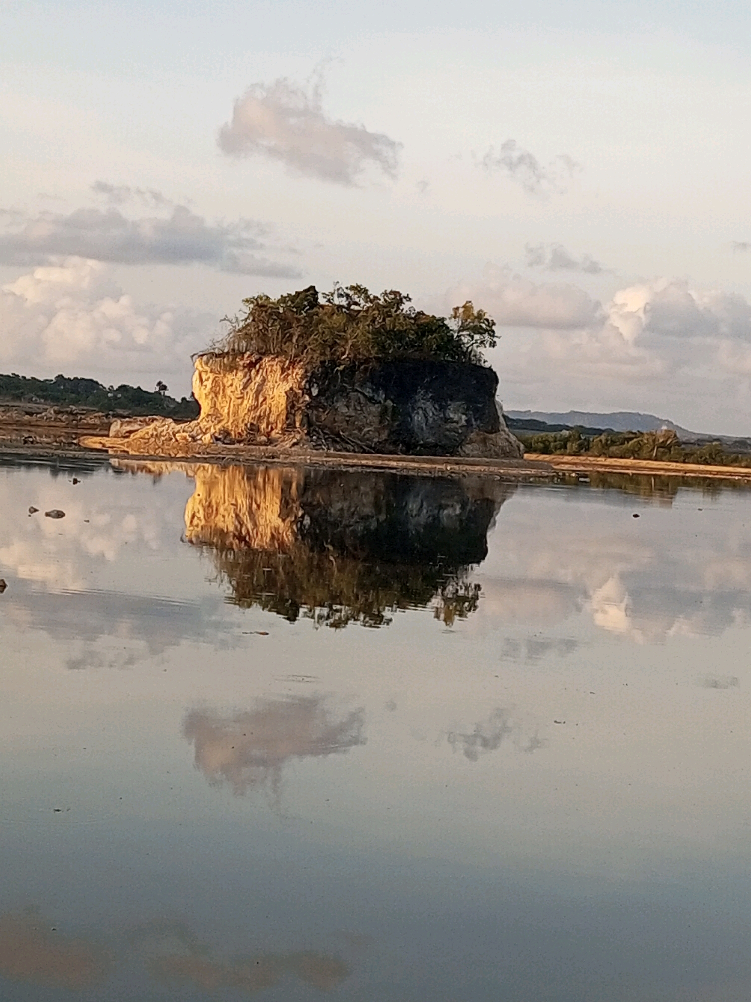 A solitary, tree-covered rock formation rises from a calm body of water, reflecting its sun-drenched facade. The rock, illuminated by warm, golden light, is textured and appears to be eroding. The water's surface is still, creating a mirror image of the rock and the overcast sky, which is dotted with soft, white clouds tinged with pinkish hues. The background shows a gently sloping, vegetated shoreline and distant hills. The scene is serene and tranquil, suggestive of a coastal or island landscape. The location is identified as Serubeba, Indonesia.