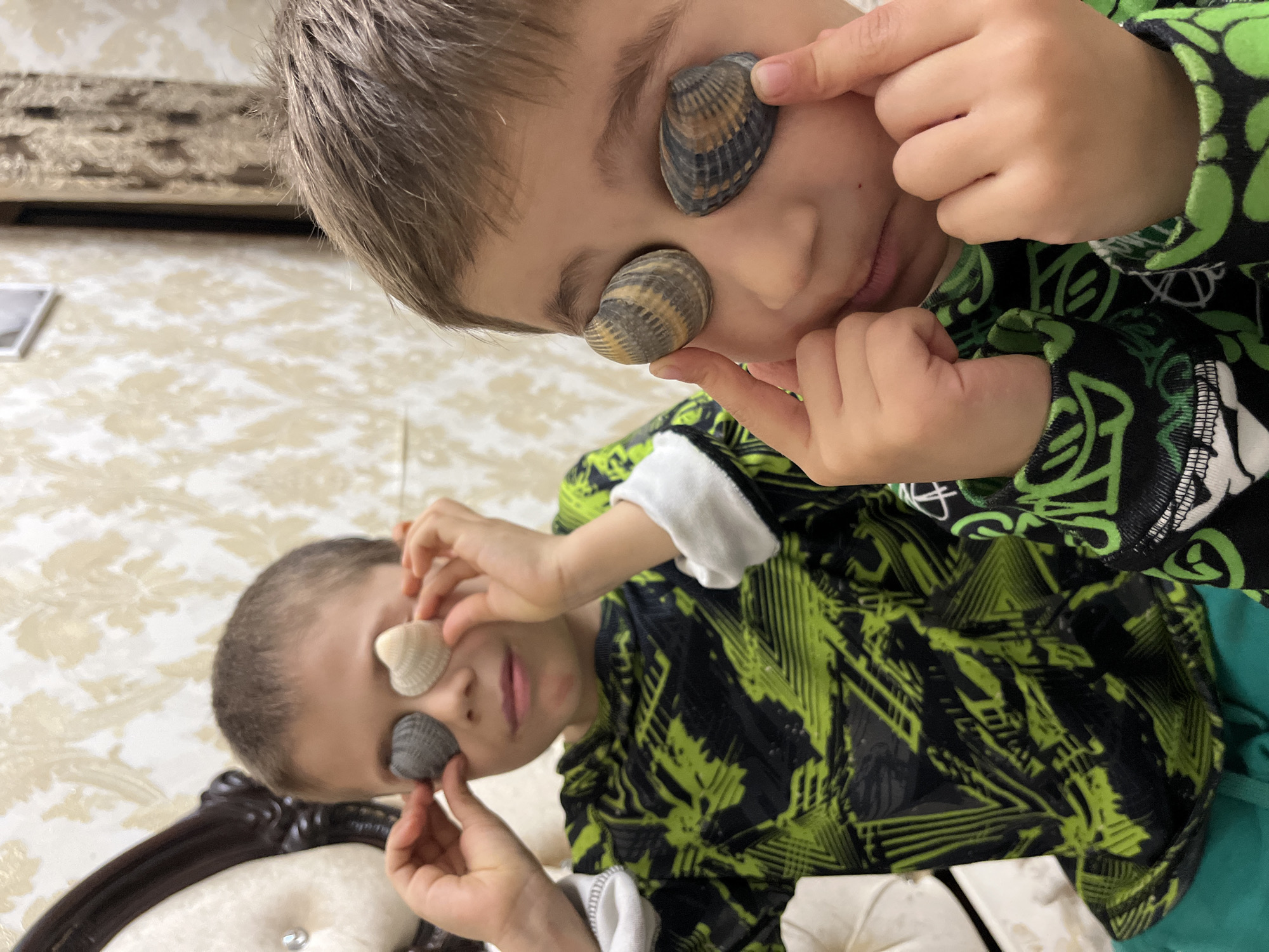 Two young boys are depicted indoors in the city of Kaspiysk, Russia, each holding seashells over their eyes. The boy in the foreground, positioned to the right, has light brown, spiky hair and wears a black long-sleeved shirt with vivid green, abstract patterns. He holds two ridged, bivalve seashells, striped in shades of grey-brown and beige, over his closed or partially closed eyes using his index fingers. His mouth is slightly open. The boy in the background, positioned to the left and slightly out of focus, has dark brown, short hair and wears a black long-sleeved shirt with sharp green geometric patterns. He holds two smaller, heart-shaped bivalve seashells over his eyes: one is dark grey, and the other is light beige. His gaze is directed downwards. Both boys have light-colored cuffs on their sleeves. The background features a light-toned wallpaper with a subtle, repeating gold or beige damask-like pattern. To the upper left, a section of a cream-colored upholstered headboard or chair with a dark carved wooden frame and a decorative clear button is visible. In the upper right background, a light-colored wall with the same wallpaper transitions into a darker, possibly curtained, opening, above which a small framed object is visible on the wall.