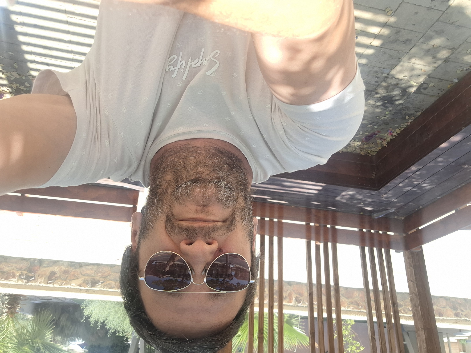 A man in sunglasses and a light-colored t-shirt is taking a selfie outdoors. He has a beard and is standing or sitting on a wooden bench in what appears to be an outdoor seating area with a slatted wooden roof. The background shows palm trees and stone walls, consistent with a location in Yerocham, Israel. The ground is paved with stone tiles, and there are fallen leaves scattered around. The t-shirt has a subtle geometric pattern and the word "Simplify" printed on it.
