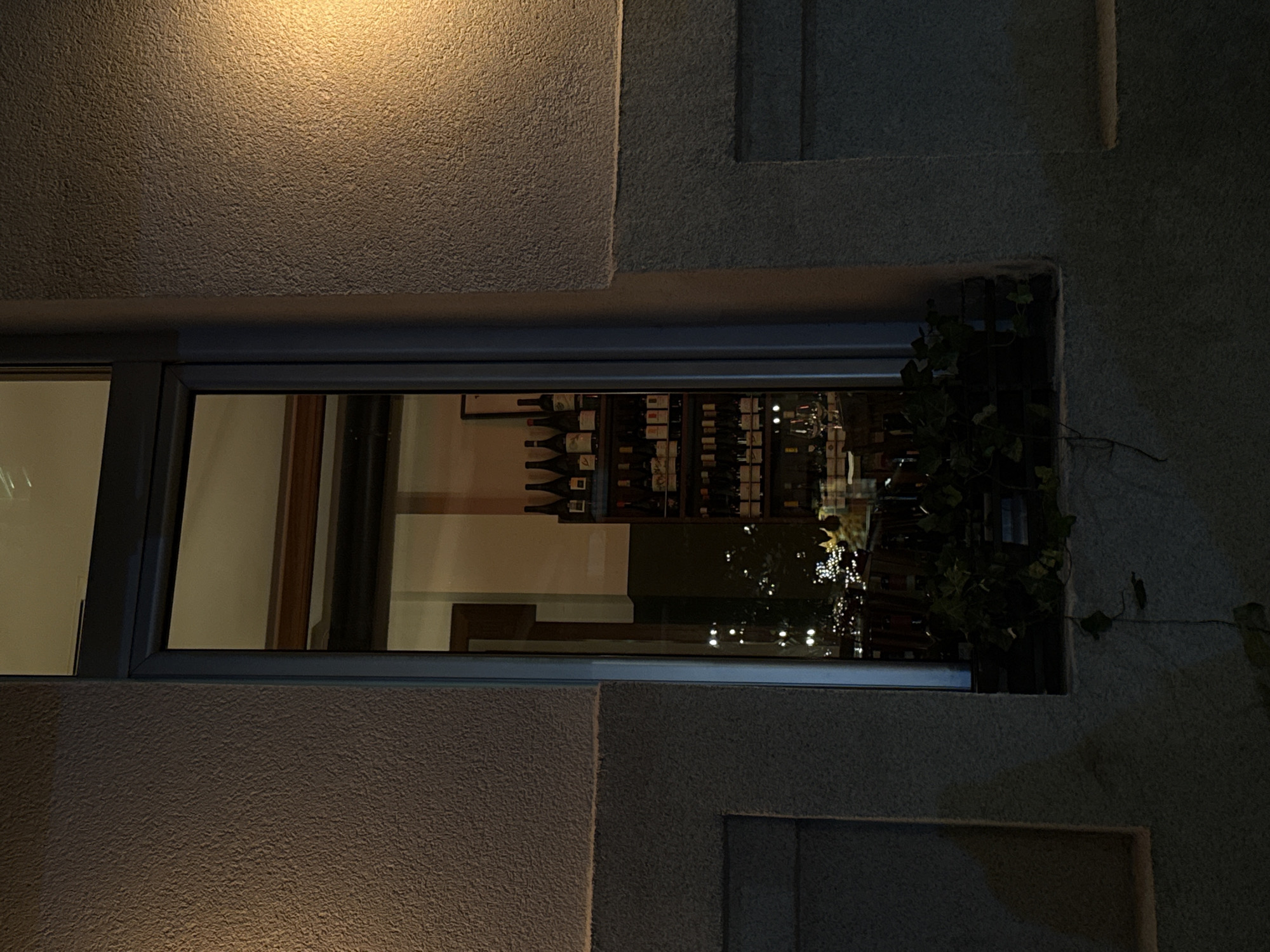 The image shows a window of a building at night. Inside the window, there is a display of wine bottles on shelves, suggesting a wine shop or restaurant. The shelves are well-stocked with various types of wine bottles, and some glassware is visible on a table in the foreground. Outside the window, there is a textured wall with some decorative elements, and a light source illuminates the wall from the right. The overall scene is dimly lit, with reflections in the window adding to the atmospheric quality. The location is identified as Belgrade, Serbia.