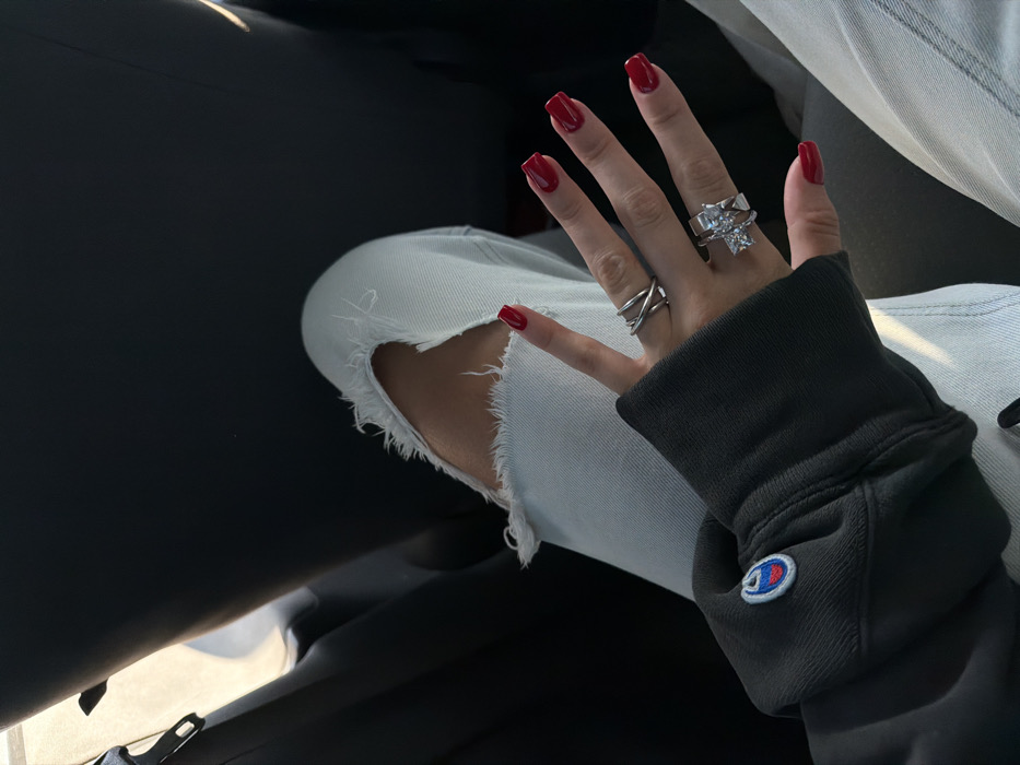 A person sits inside a car, knee bent and facing the camera. The person wears distressed light wash denim jeans and a dark gray Champion sweatshirt. The hand is visible and adorned with two silver rings. The nails are painted a shiny red color. The background is dark and blurry, with the car interior partially visible. The scene is softly lit, highlighting the texture of the denim and the rings.