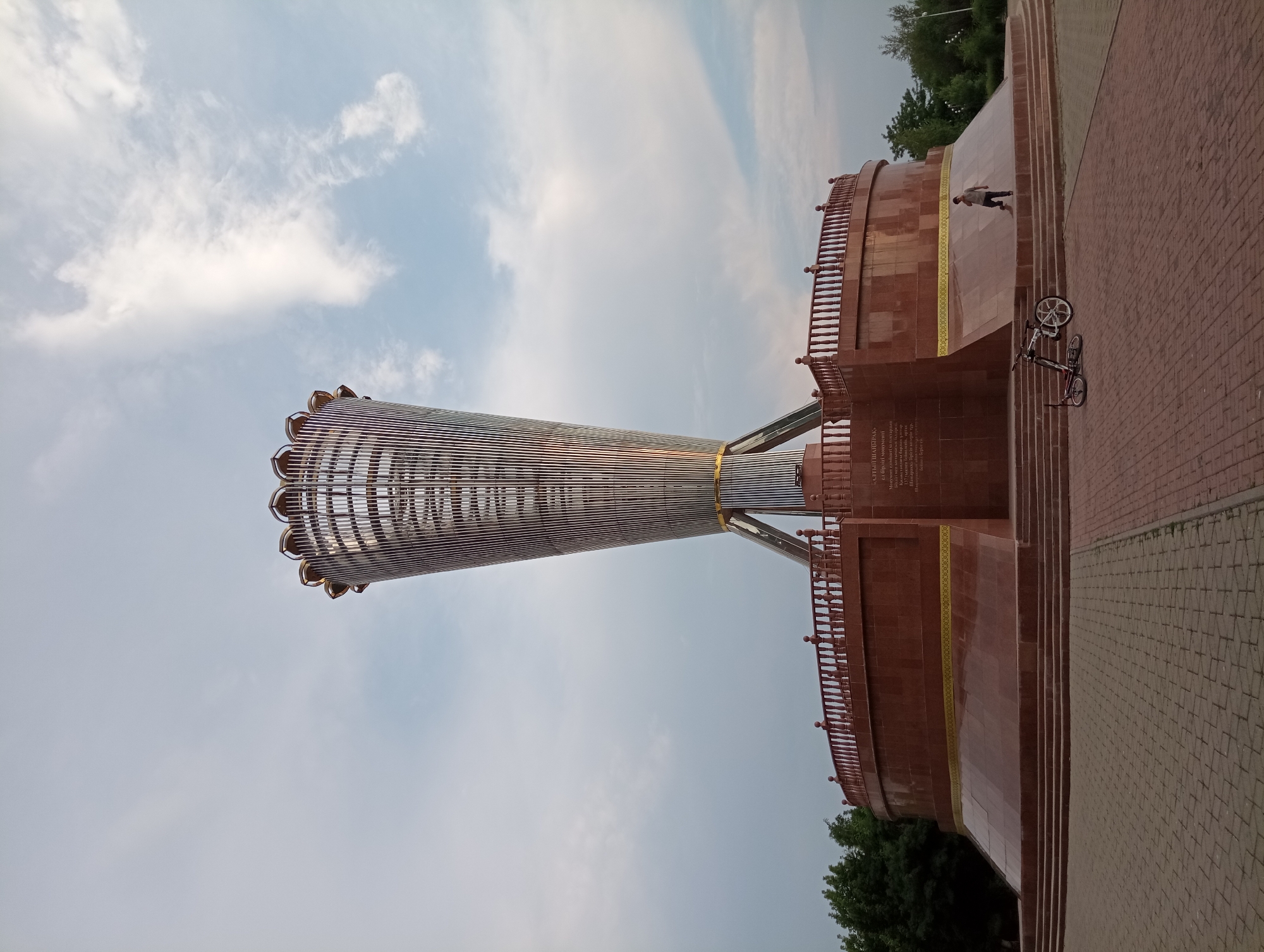 The image shows the Monument of Independence in Shymkent, Kazakhstan. The monument is a tall, slender structure with a metallic, ribbed design that tapers towards the top, resembling an opened flower or a torch. It stands on a large, multi-tiered circular base made of reddish-brown stone, with a decorative railing around the top level. The base also features engraved text.
In the foreground, on the paved area leading up to the monument, are two bicycles. One is standing upright, and the other is lying on its side. A lone individual, wearing a grey shirt and dark pants, is walking on the side of the monument's base. The surrounding environment includes green trees to the left and right of the monument, and a cloudy sky above. The overall atmosphere suggests an outdoor public space, possibly a park or square.
