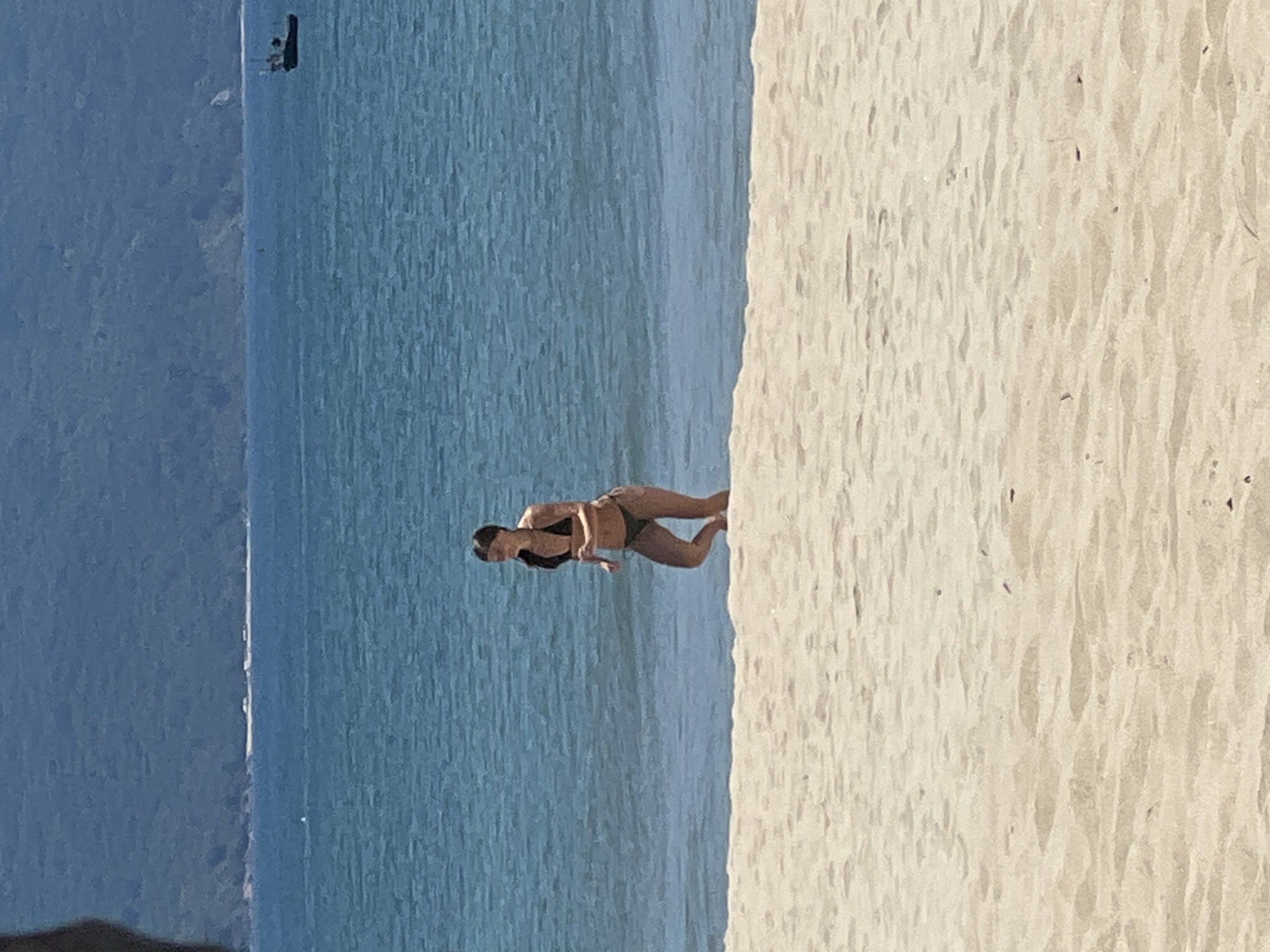 The image shows a young woman running on the shallow water at the edge of a sandy beach. She is wearing a dark bikini and her hair is tied back. The water is a clear turquoise blue and the sand is light colored. In the background, there is a large body of blue water, a distant green mountain range, and a small boat on the water. The scene appears to be a tropical beach, likely in a location like Đà Nẵng, Vietnam, known for its long sandy beaches and clear waters. The lighting suggests a sunny day.