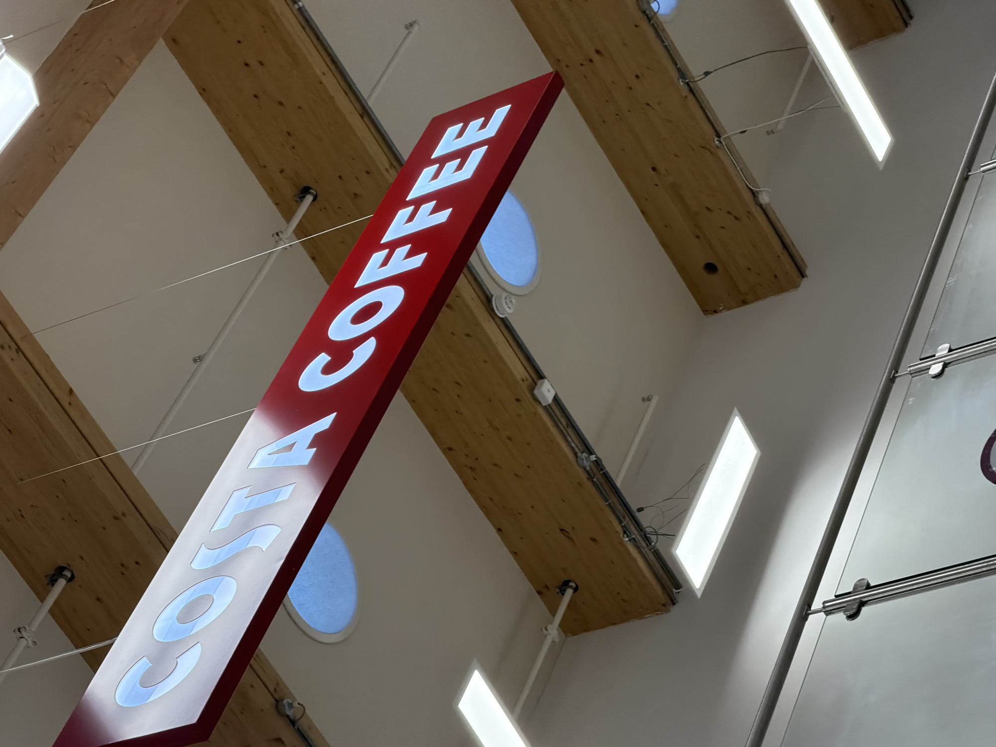 A tilted, upward-angled shot captures a red Costa Coffee sign suspended from a high ceiling. The white lettering of "COSTA COFFEE" is illuminated. The ceiling features a combination of bare wooden beams and smooth white panels with round windows. Linear lighting fixtures are also visible, casting bright light onto the scene. Part of a glass structure with metal supports is visible on the right side of the image. The location is suggested to be Pity Me, United Kingdom.