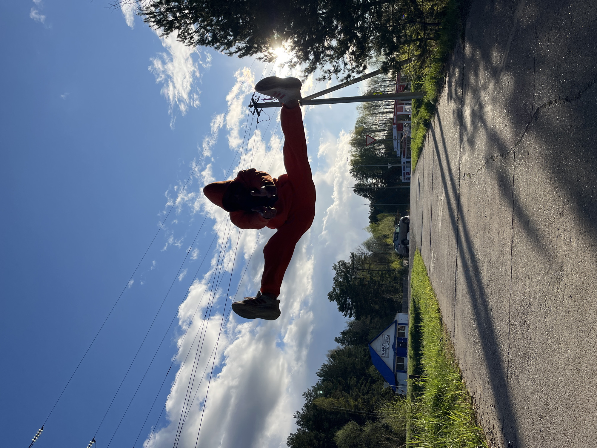 A person wearing an orange tracksuit is standing upside down with one leg extended upward and one leg extended downward. Their face is not visible. They are positioned near power lines against a backdrop of a blue sky with scattered white clouds. In the background is a road, trees, and a building with the word "KAPE" visible, suggesting the location is Chaikovsky, Russia.