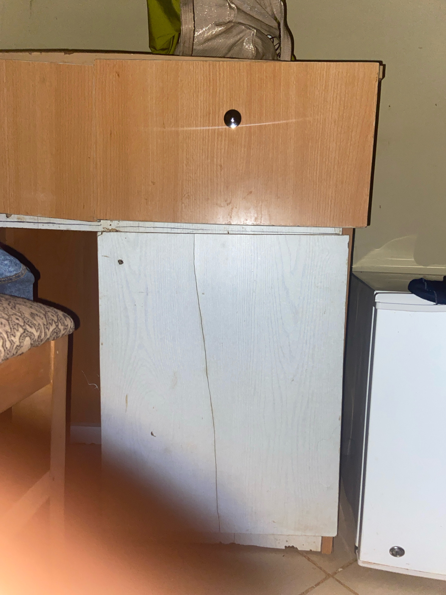 A close-up shot shows the front of a light-colored, wooden dresser or cabinet with a slightly darker wooden top. A circular, metallic handle is centered on the top drawer of the dresser. Below the top drawer is another section of the cabinet, which appears to be a door or a drawer front, made of a lighter material, possibly white laminate, with a visible crack running vertically down the center. To the right of the dresser is a white appliance, likely a refrigerator or freezer, with a similar metallic handle visible on its front. To the left of the dresser, a portion of a bed with a patterned pillow and blue fabric is visible. The environment appears to be indoors, with a tiled floor and a light-colored wall in the background. The overall impression is one of a cluttered, lived-in space.