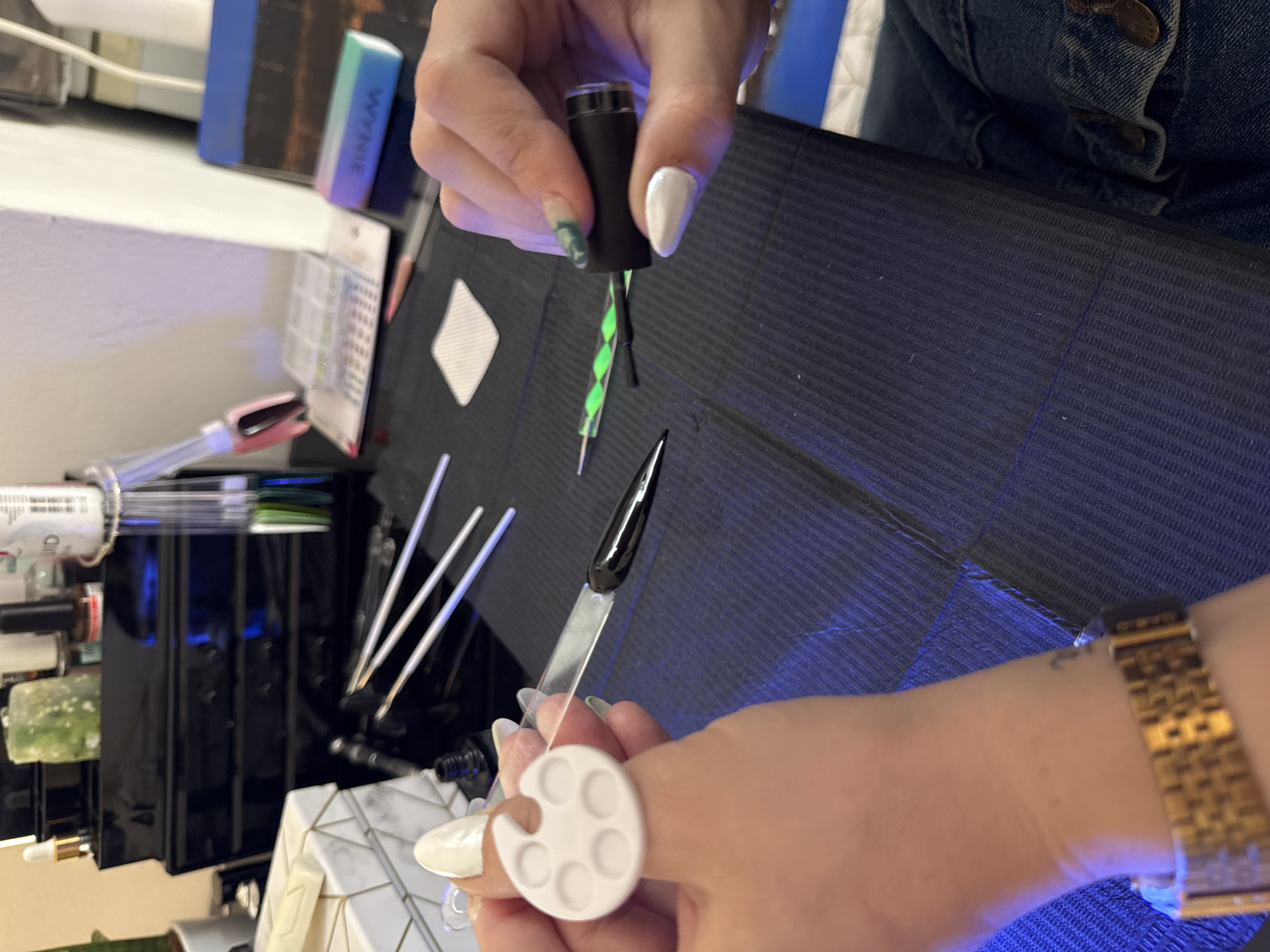 The image depicts a close-up view of a manicure being performed, possibly at a nail salon or private residence. A person is holding a black nail polish bottle with a green and black brush, applying black polish to a pointed artificial nail tip held on a white plastic stick with a white palette ring. Several white-handled brushes are visible in the background, along with other nail care products and a blue and green nail file. The background consists of a black textured surface and what seems like a container or cabinet. A person wearing a gold watch is visible on the right side of the image. The location and the city of Yaiza, Spain, are not verifiable from the image provided.