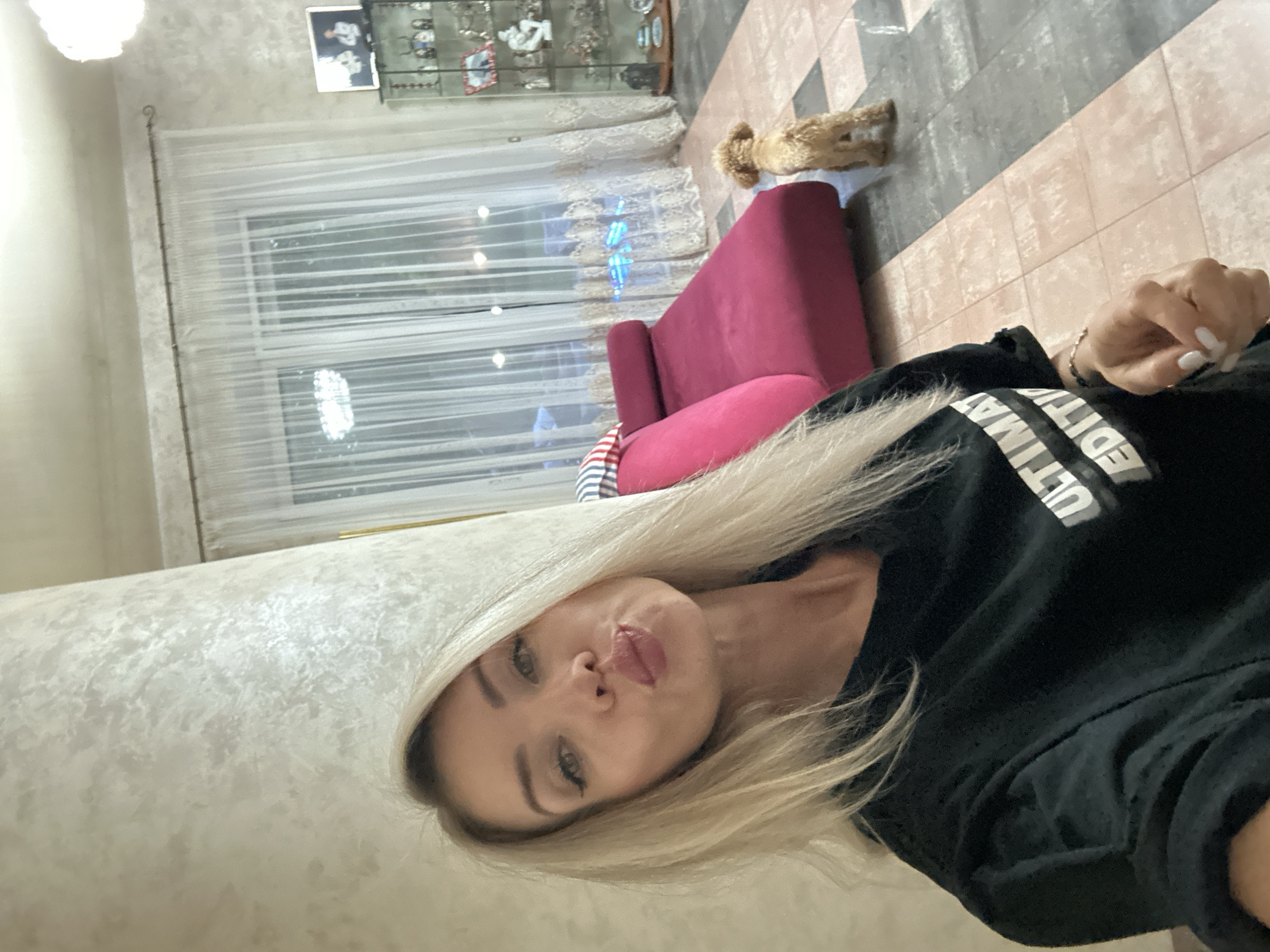 A blonde woman with fair skin, wearing a black t-shirt with white text, is taking a selfie. She has fair skin, light brown eyes, and is wearing makeup. Her lips are pursed as if blowing a kiss. In the background, a small tan dog stands on a tiled floor. A bright pink chaise lounge is visible behind the dog. The room appears to be a living area with a large window covered by sheer white curtains. A tall glass display cabinet filled with decorative items is to the right of the window. The walls are textured, and the floor is a mix of light pink and gray tiles. There are no identifying markers in the image to confirm its location in Armavir, Russia.