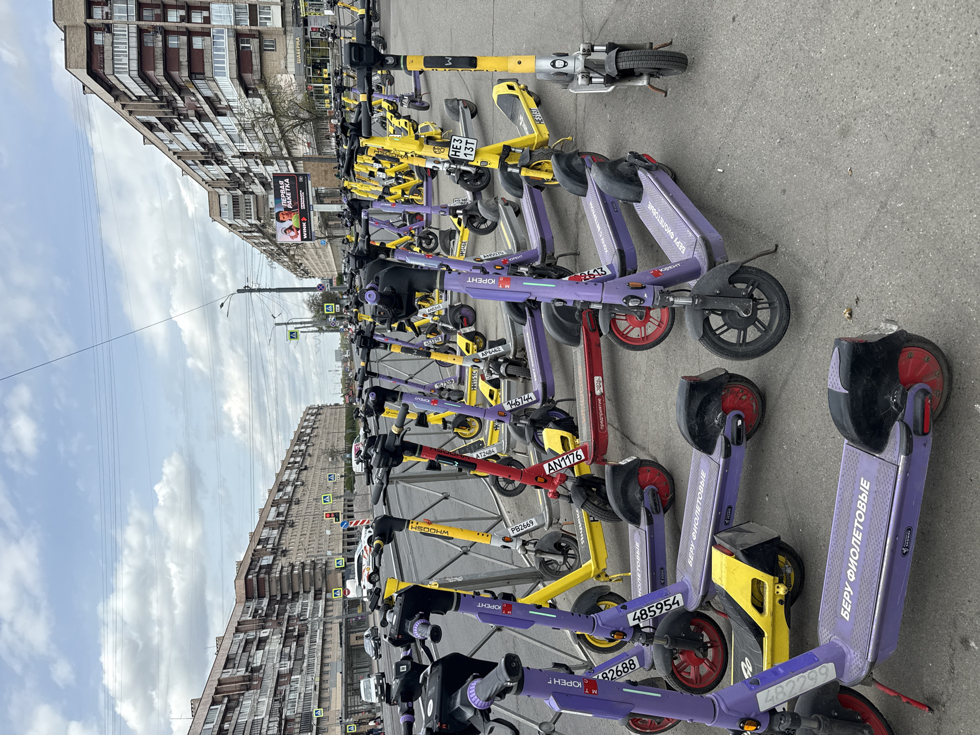 The image shows a row of parked electric scooters on a city street. The scooters are a mix of purple and yellow colors, with license plates. There are multiple buildings on both sides of the street, and a blue sky with clouds. A large advertisement board is also visible on one of the buildings. Based on the signage in Russian and the scooter brand identified as "Yurent", the location is likely to be in a city in Russia, and possibly Saint Petersburg.
