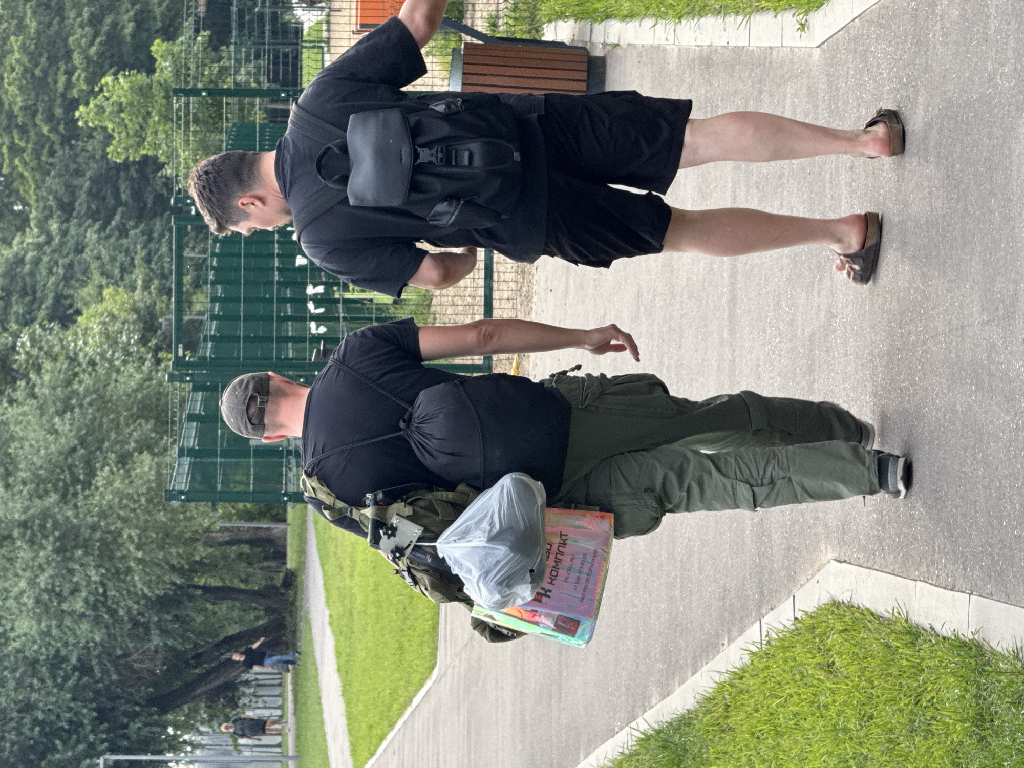 Two men walk away from the camera on a paved pathway in an outdoor setting. The man on the left wears a black t-shirt, green cargo pants, and a baseball cap. He carries a large olive green backpack with a smaller black drawstring bag attached. A plastic bag containing an item and a holographic shopping bag with Cyrillic text and a phone number are also attached to his backpack. The man on the right wears a black t-shirt, black shorts, and dark sandals. He carries a black backpack. In the background, a green fence, trees, and grass are visible. Distant figures can be seen walking on a grassy area to the left. The environment appears to be a park or a recreational area, possibly within the city of Moscow, Russia, given the signage and the general urban park aesthetic.