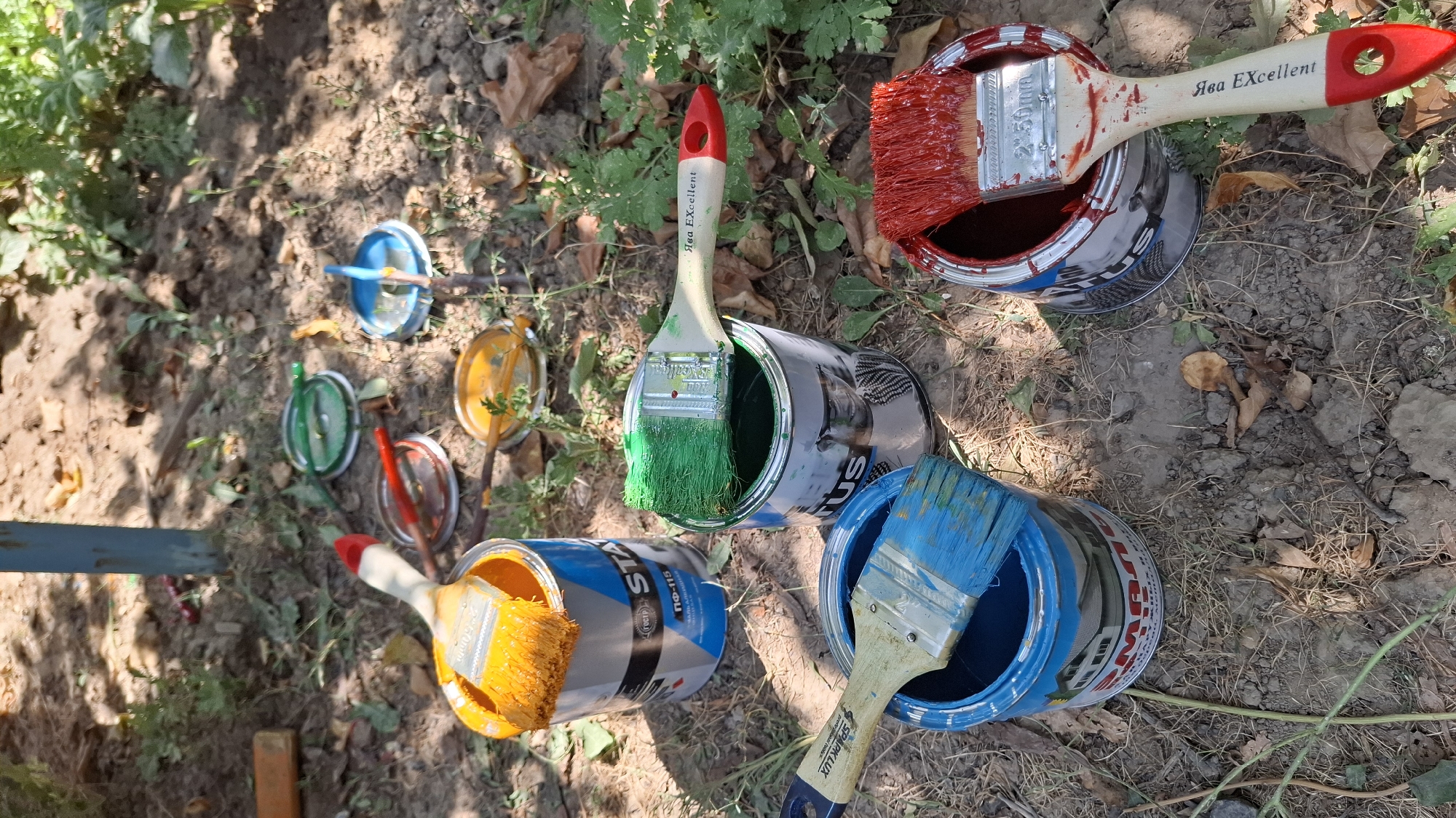 In Шұбарсу, Kazakhstan, an outdoor scene depicts several cans of paint and paintbrushes on the ground. The environment is a mix of dirt, dry grass, and some green foliage, suggesting a garden or yard area. Sunlight casts shadows, indicating a bright day.
There are at least four open cans of paint, each with a different color: orange, green, blue, and red. Each can is accompanied by a paintbrush resting on its rim or inside, with bristles loaded with paint.
The orange paint can has a brush with orange paint. The green paint can has a brush with vibrant green paint, and the handle of this brush has text that appears to be "Ява Excellent". The blue paint can holds a brush with blue paint. The red paint can contains red paint, and the brush placed on its rim also appears to have red paint on its bristles. The handles of the brushes are made of light-colored wood with red accents on some.
In the background, several colored lids are scattered on the ground, corresponding to the colors of the paint cans: green, red, orange, and blue. A blue metal pole is partially visible in the upper left corner of the image. The overall impression is that painting work is in progress or has recently occurred outdoors.