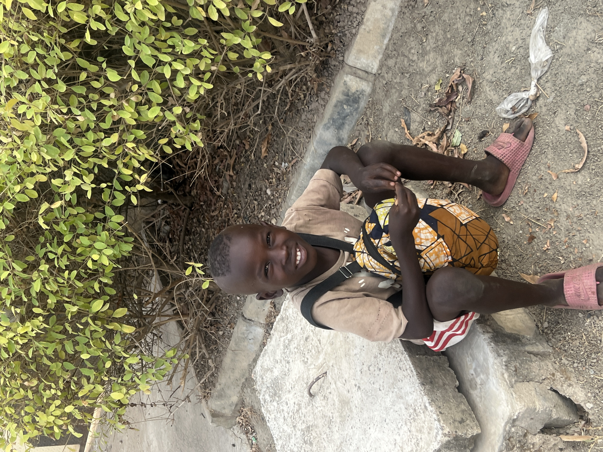 A young boy sits on a concrete block in Minna, Nigeria. He is dark-skinned, with short hair, wearing a beige t-shirt and red and white striped shorts. He has a satchel-like bag on his shoulder. He wears pink flip-flops. He smiles, looking up at the camera. Beside him is a green leafy bush. The ground is dirt and gravel with some trash.