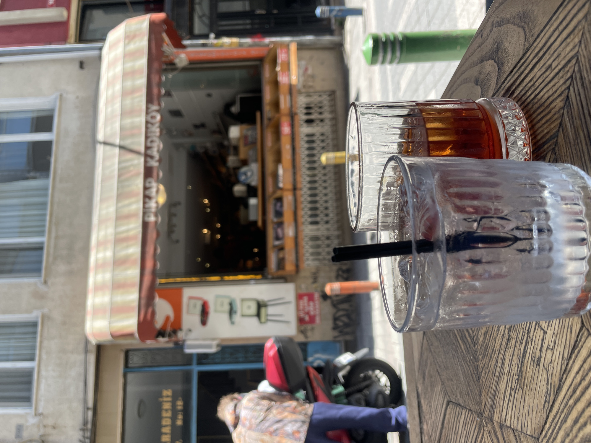 The scene is an outdoor cafe in Kadıköy, Turkey. A table with a herringbone-patterned wooden surface is the primary focus. Two glasses sit on the table: one filled with a reddish-brown liquid, possibly tea, and the other with a clear liquid and a black straw. In the background, a shopfront with a striped awning reads "PIKAP KADIKÖY." Inside the shop, shelves are visible. Outside, a motorcycle is parked, partially obscured by a person wearing a patterned shirt and blue pants. The street setting includes buildings and a sidewalk.