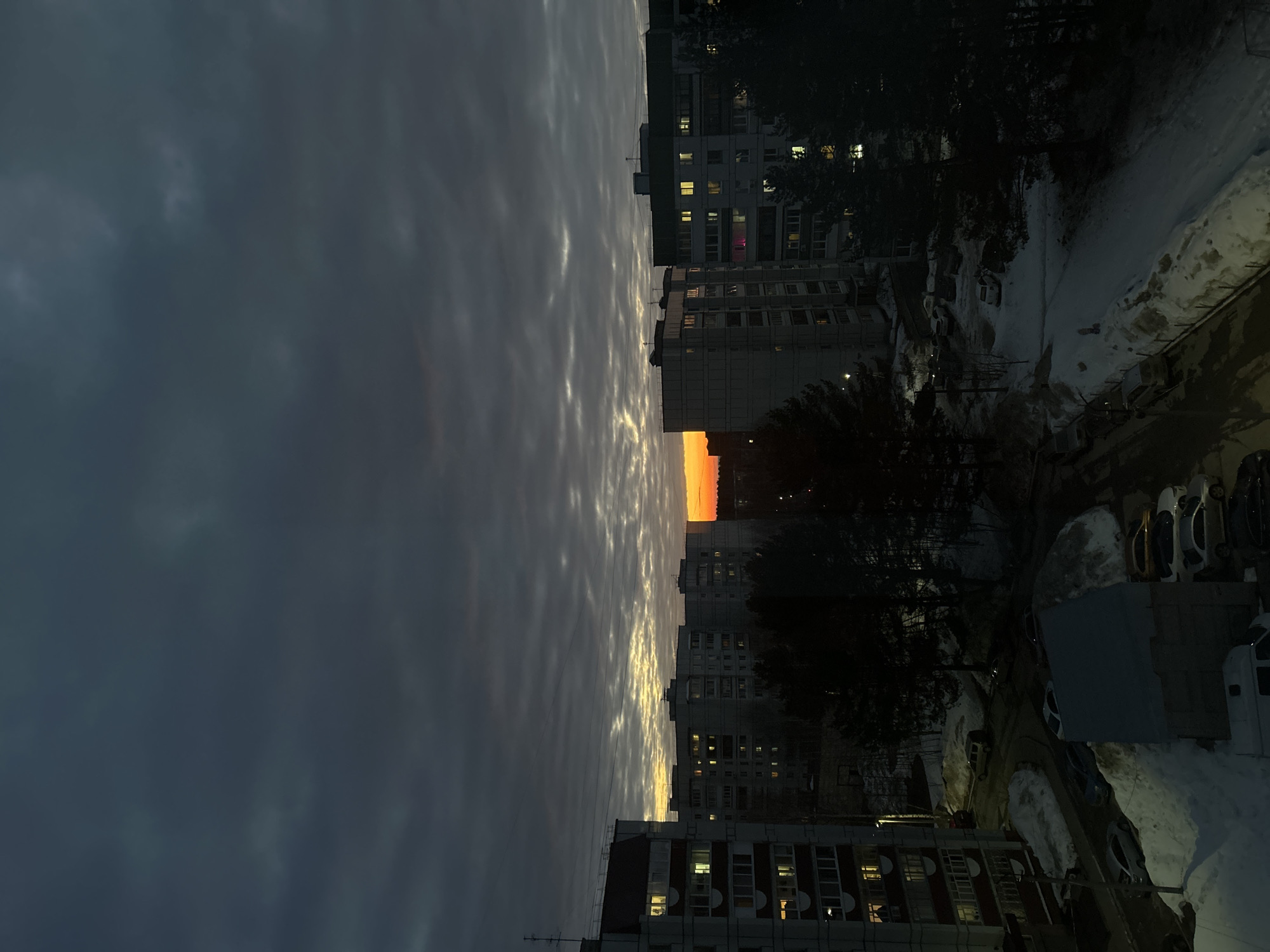 The image displays an urban landscape in Chaikovsky, Russia, observed from an elevated perspective during either sunrise or sunset. The sky is dominated by dark grey altocumulus clouds, with a distinct horizontal band of bright orange light visible on the horizon, indicating the sun's position.
Several multi-story residential buildings, characteristic of Soviet-era architecture with light-colored facades and numerous windows, populate the midground and background. Many windows in these structures exhibit warm yellow illumination, suggesting interior lighting is active. A prominent apartment building occupies the left foreground, featuring a visible rooftop antenna and multiple lit windows.
The ground throughout the scene is covered in snow, indicating cold weather conditions. Snowdrifts are visible along the edges of paved areas. Dark, coniferous trees, likely pines or spruces, are interspersed between the buildings and line roads, their foliage largely unlit and appearing as silhouettes against the snow and ambient light. Paved roads and parking areas are present, with several parked vehicles visible on the snow-dusted surfaces. A single-story structure with a dark roof is located in the bottom center. No individuals, explicit actions, or interactions are discernible within the frame.