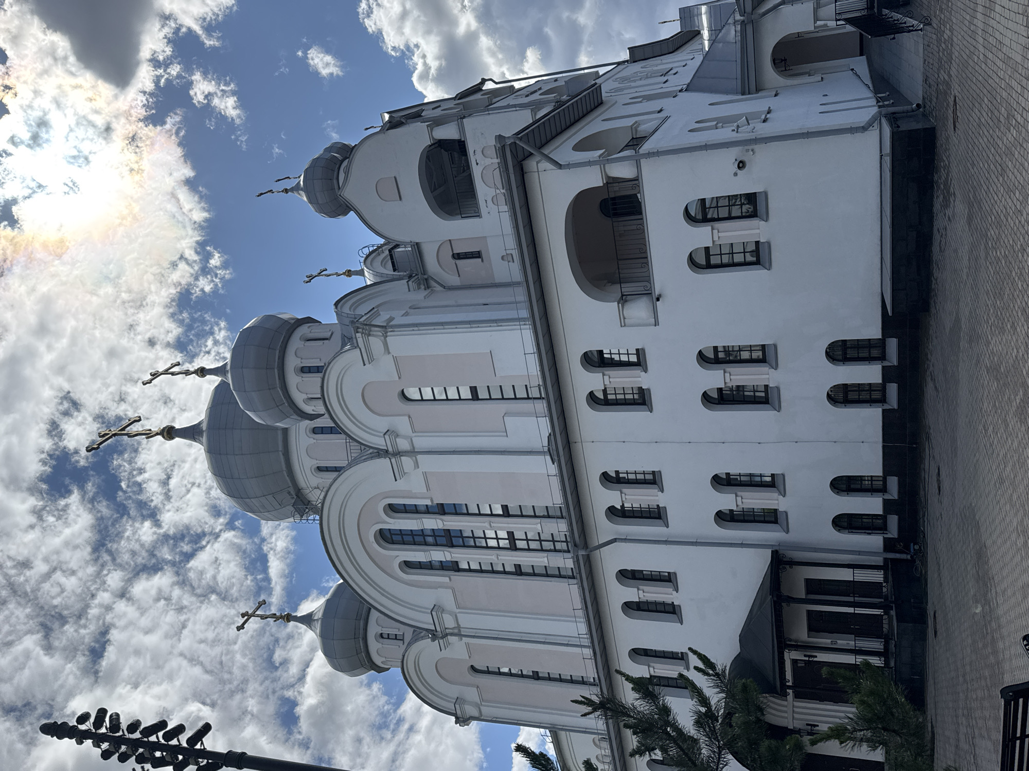 The image shows the Church of the Kazan Icon of the Mother of God in Saint Petersburg, Russia. The church is a large, white, domed building with a pale pink trim. It has numerous arched windows and balconies. The sky is blue with white clouds, and the sun is visible in the upper right corner, casting a rainbow-like effect through the clouds. A street light is visible on the left side of the image. The ground in front of the church is paved with grey bricks. There are no people visible in the image.