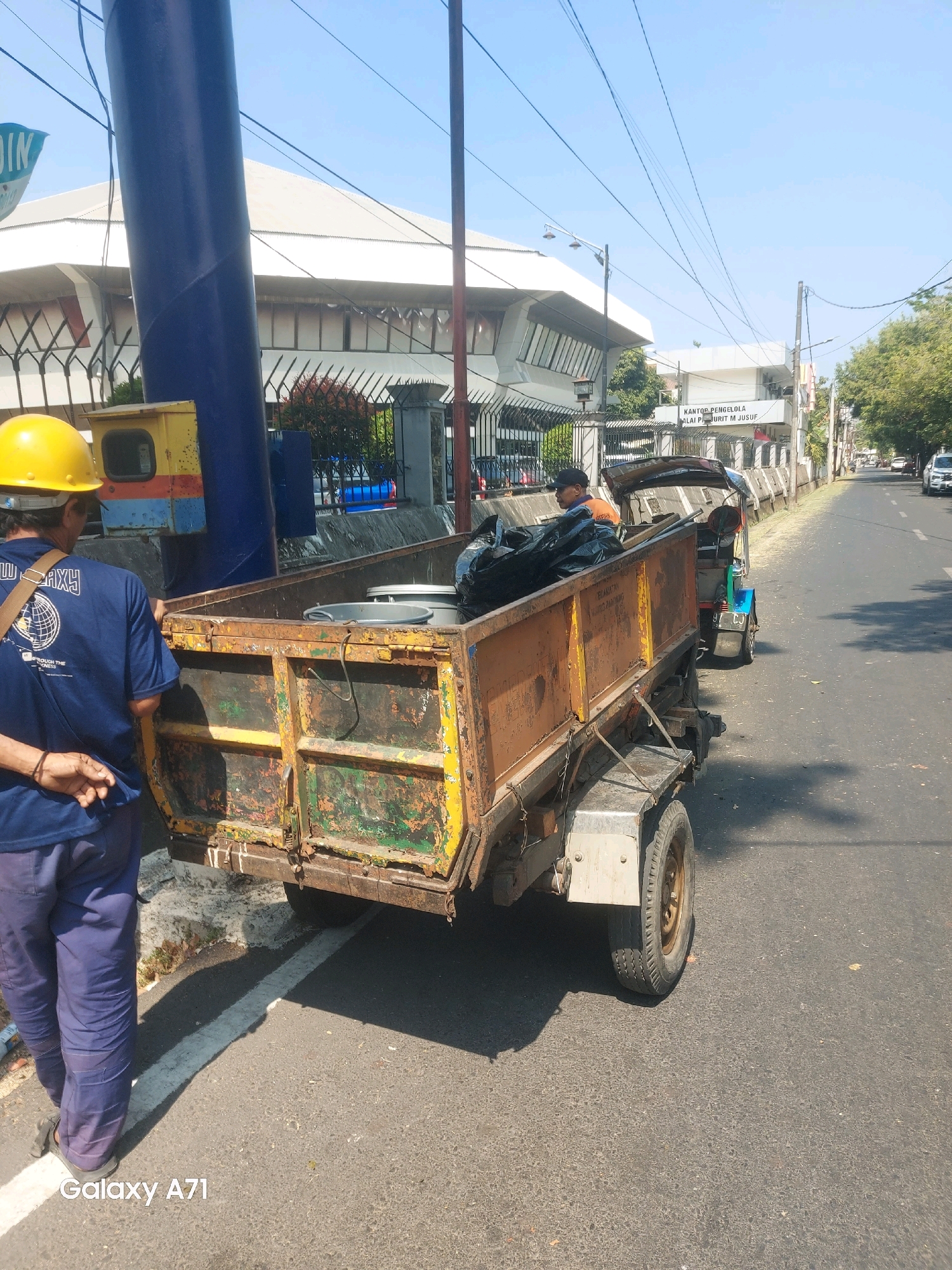 In Makassar, Indonesia, a man wearing a yellow hard hat and a blue shirt with "GALAXY" printed on the back stands beside a dilapidated orange and green trailer attached to a small motorized vehicle, possibly a trishaw. The trailer is filled with black garbage bags and some metal buckets. In the background, a white building with a sloped roof is visible, along with a blue pole and a sign that reads "DIN". Further down the street, another building has a sign that says "KANTOR PENGELOLA ALAT PRIBADI M JUSUF." The scene is set on a sunny day, with power lines crisscrossing the sky.