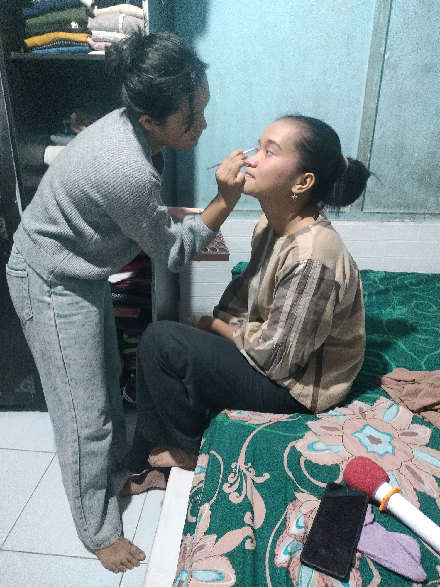 In a dimly lit room, likely a bedroom in Pekanbaru, Indonesia, a young woman is applying makeup to another young woman. The woman applying makeup, dressed in a grey knitted sweater and light-wash jeans, stands and leans forward, holding a makeup brush to the face of the seated woman. The seated woman, wearing a beige and brown striped shirt and dark pants, sits on a bed covered with a green floral-patterned sheet. Her hair is pulled back in a bun, and she wears small earrings. On the bed next to her, a black smartphone, a red-capped microphone, and a purple cloth are visible. In the background, a dark wardrobe is filled with folded clothes. The floor is tiled.