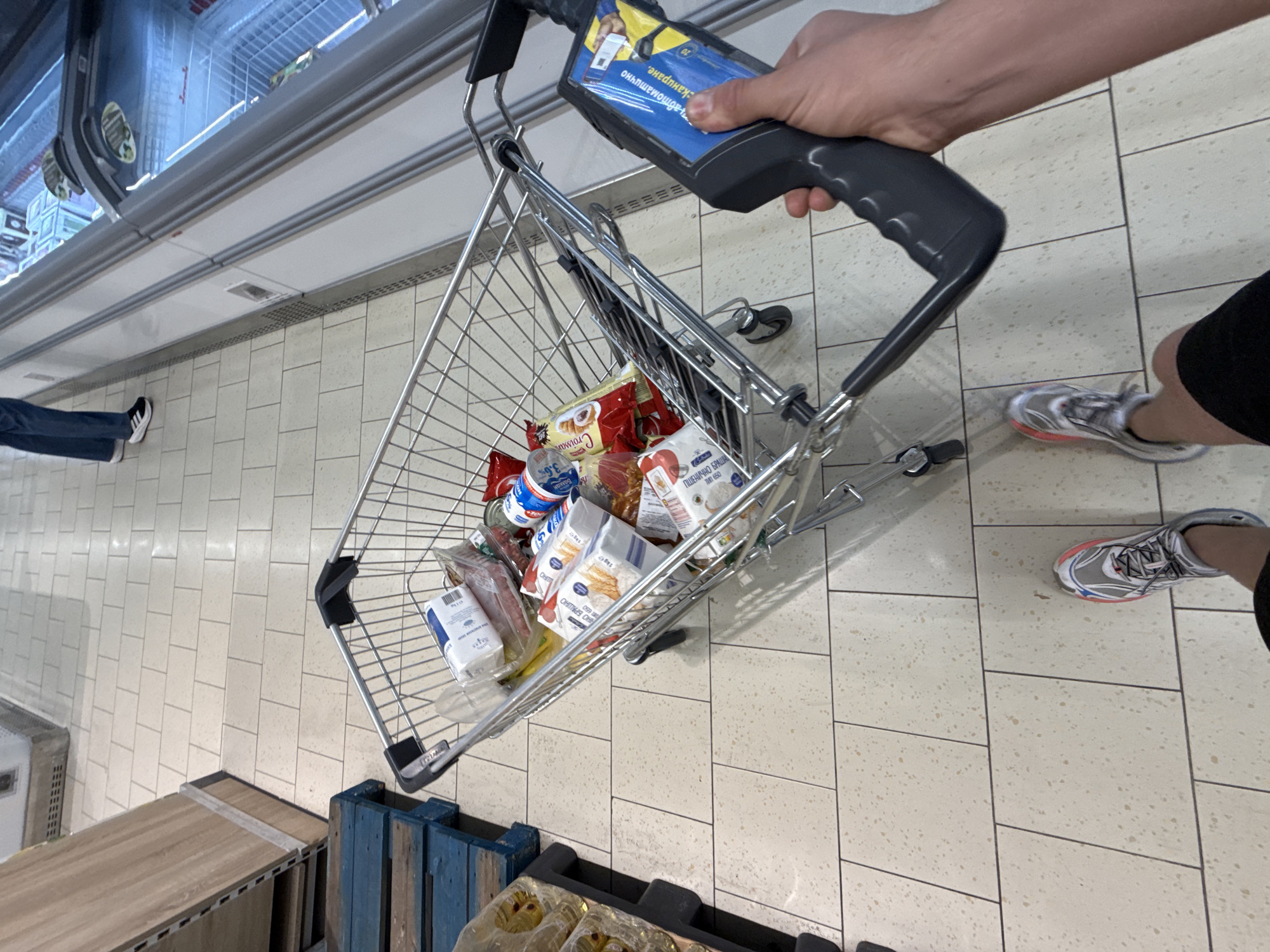 A first-person perspective shot captures a shopping cart filled with groceries and a scanner in the right hand of the person holding the camera. The shopping cart contains various food items, including bags of flour, what appears to be meat, and a carton of milk. The scanner is black with a blue and yellow sticker featuring text in Cyrillic script. In the background, a person's legs are visible wearing blue jeans and white sneakers, standing next to a refrigerator unit in what appears to be a supermarket. The floor is tiled with a light-colored pattern. The scene is likely in a supermarket in Stara Zagora, Bulgaria, given the Cyrillic text on the scanner and the general appearance of a European supermarket.