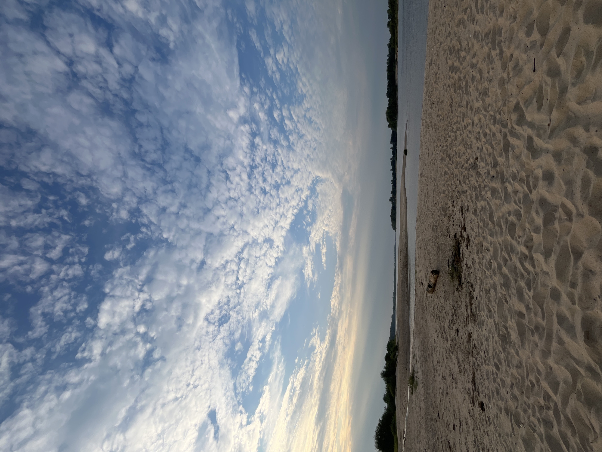 The image depicts a tranquil scene of a sandy riverbank with a wide expanse of water and a cloudy sky. The foreground is dominated by textured sand, showing ripples and footprints, suggesting a recent receding tide or wind action. A single piece of driftwood lies on the sand in the middle ground, adding a natural element to the composition. Beyond the sand, a calm river stretches across the frame, with lush green trees lining the opposite bank. The sky is a dramatic canvas of blue interspersed with numerous white, puffy clouds, some of which are illuminated by soft sunlight. The overall atmosphere is serene and natural, typical of a riverside landscape. The location is described as the city of Nizhny Novgorod, Russia.