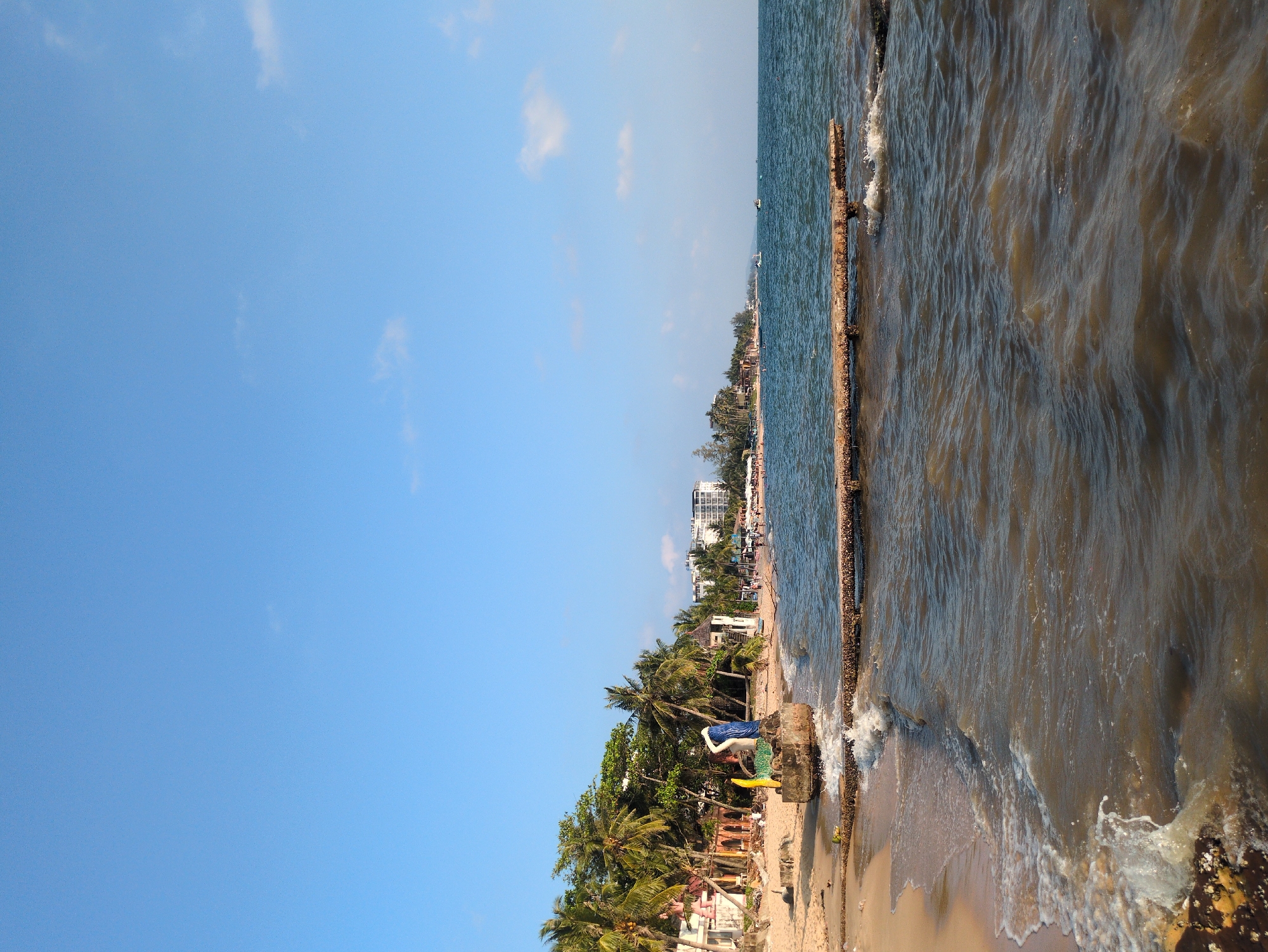 The image shows a shoreline in Unknown, Unknown. The view is taken from the beach looking towards the ocean. The sky is mostly clear blue with scattered clouds. The water is a murky brown color. A long, weathered structure, possibly a pier or seawall, runs along the edge of the water. Buildings and vegetation line the shoreline.