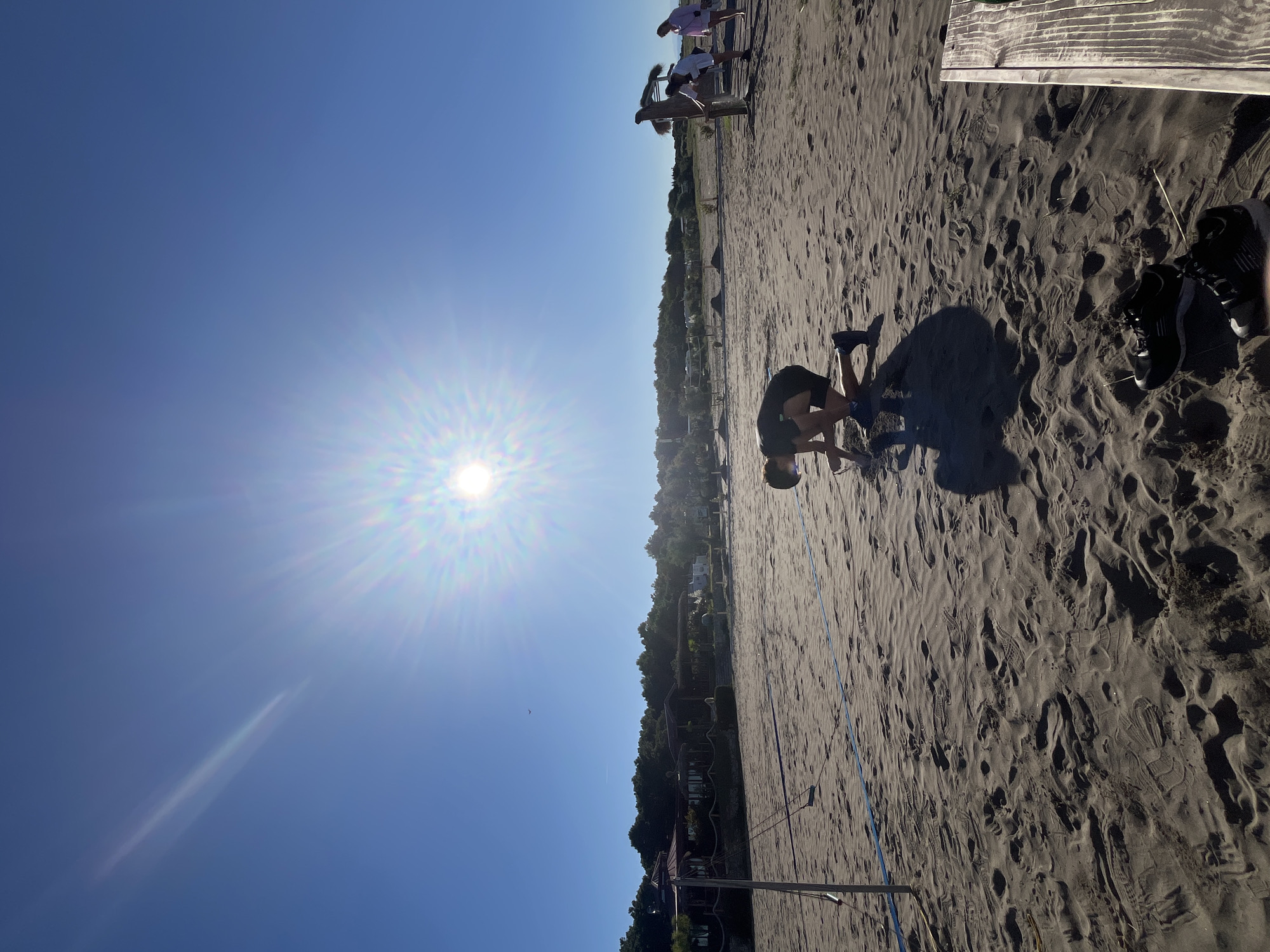 In Pistula, Montenegro, a beach scene unfolds under a bright, sunlit sky. The foreground is dominated by dark sand, marked with numerous footprints, with a pair of black athletic shoes resting on the sand to the right. A wooden bench is partially visible on the far right.
In the mid-ground, a young person in athletic wear is crouched low on the sand, appearing to be digging or examining something. Further back, a volleyball net stretches across the sand, with a single blue rope or line visible.
In the background, several structures resembling beach huts or small buildings are nestled amongst trees. To the right, three individuals are gathered around a wooden structure, possibly for exercise or leisure. The sky is a clear, vibrant blue, with the sun prominently displayed, casting a strong glare.