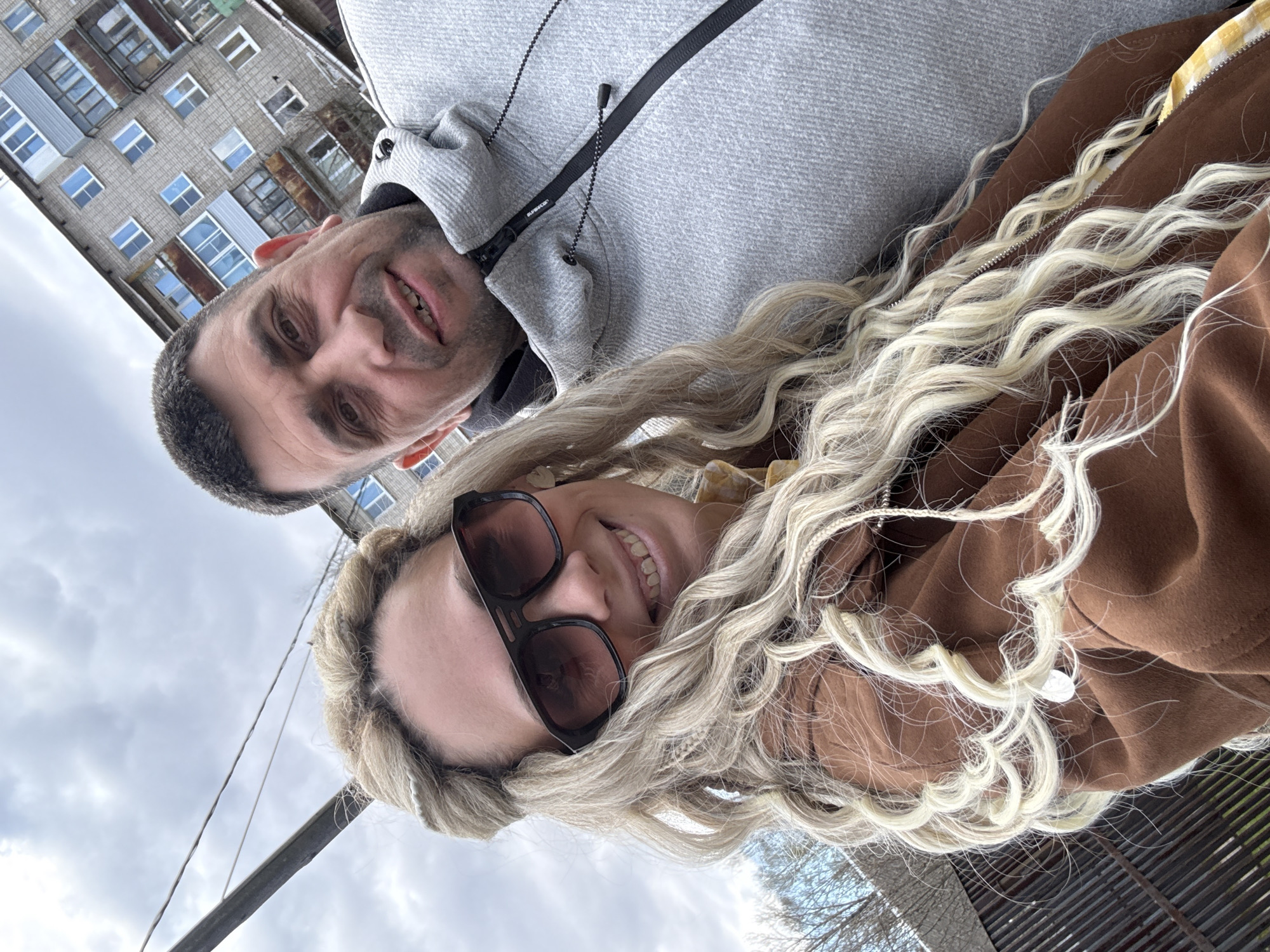 This is an outdoor selfie featuring a man and a woman, captured from a low angle looking upwards. The overall setting suggests a cloudy daytime.
The man, positioned on the left side of the frame, appears middle-aged with short, dark hair and some stubble. He is smiling, showing his teeth. He wears a grey hooded jacket, and on one of the zipper pulls, the brand "SUPERDRY" is visible. Behind him, a grey, multi-story apartment building with numerous windows, some appearing old, boarded up, or with rusty frames, stands under an overcast sky.
The woman, on the right, has long, wavy blonde hair. She is wearing dark sunglasses and is also smiling broadly, revealing her teeth. She wears a brown jacket, which has a yellow patterned lining or collar visible near the zipper.
The activity taking place is a self-portrait, possibly of a couple, expressing happiness or contentment. The background features a large building typical of urban or semi-urban residential areas, along with visible wires or cables crossing the sky. The cloudy sky is prominent. The location context is Rusanovo, Russia.